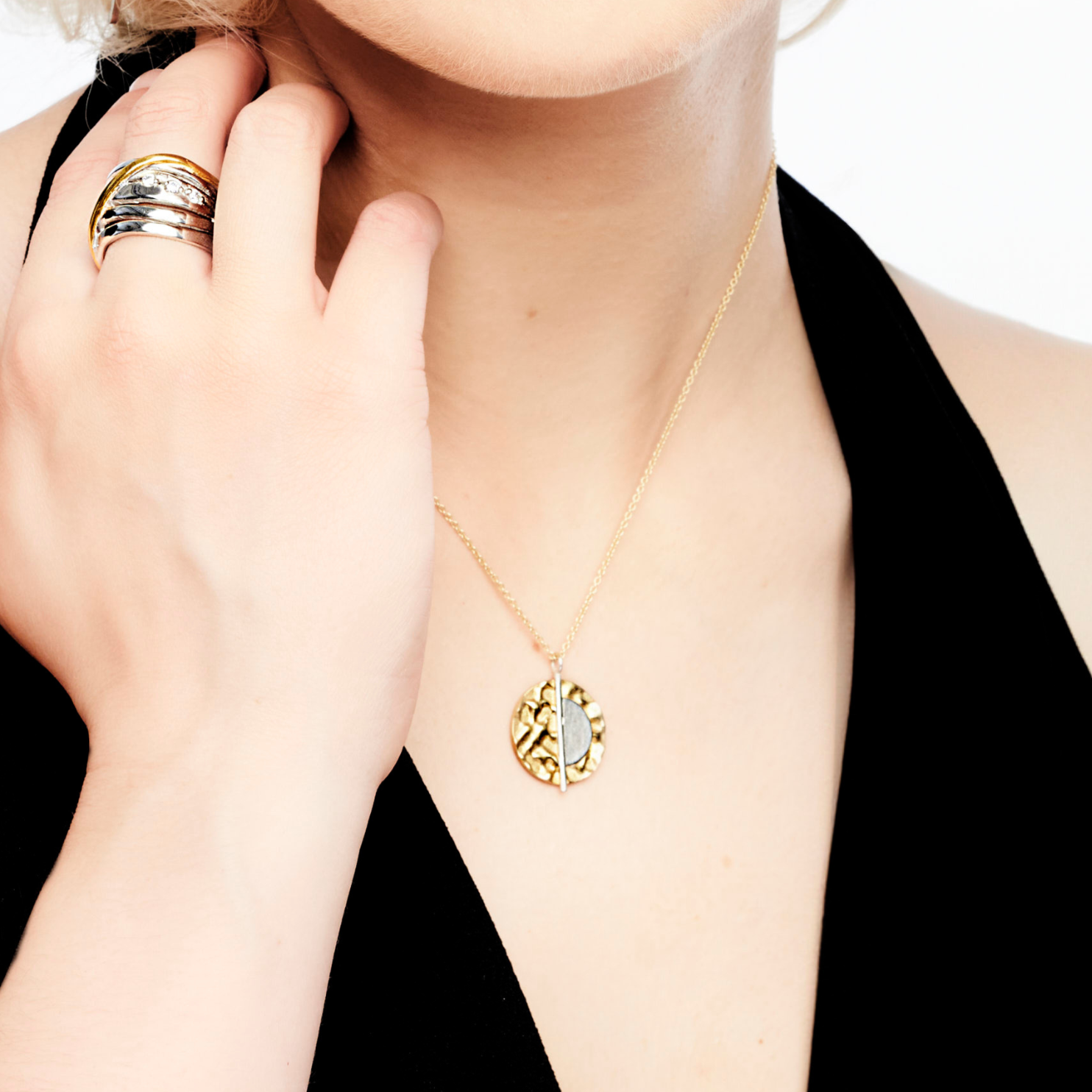A person with light skin wearing a black sleeveless top holds their chin with their left hand, showcasing a sterling silver and gold vermeil ring on their index finger. The person also wears Gallardo & Blaine Designs' Lunar Eclipse Pendant & Chain in gold