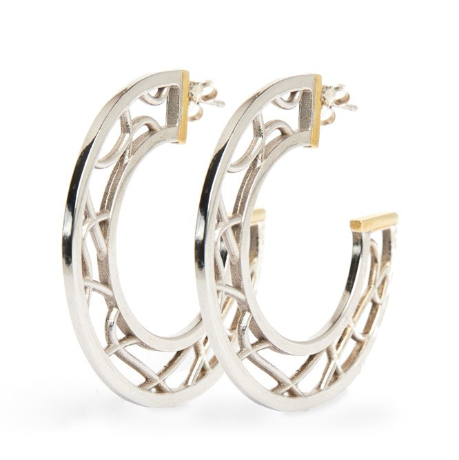 The Maya Earrings by Gallardo & Blaine Designs are a pair of intricately designed sterling silver hoop earrings with gold vermeil accents. The hoops feature open latticework in a geometric pattern, combining modern and classic elements, and are positioned
