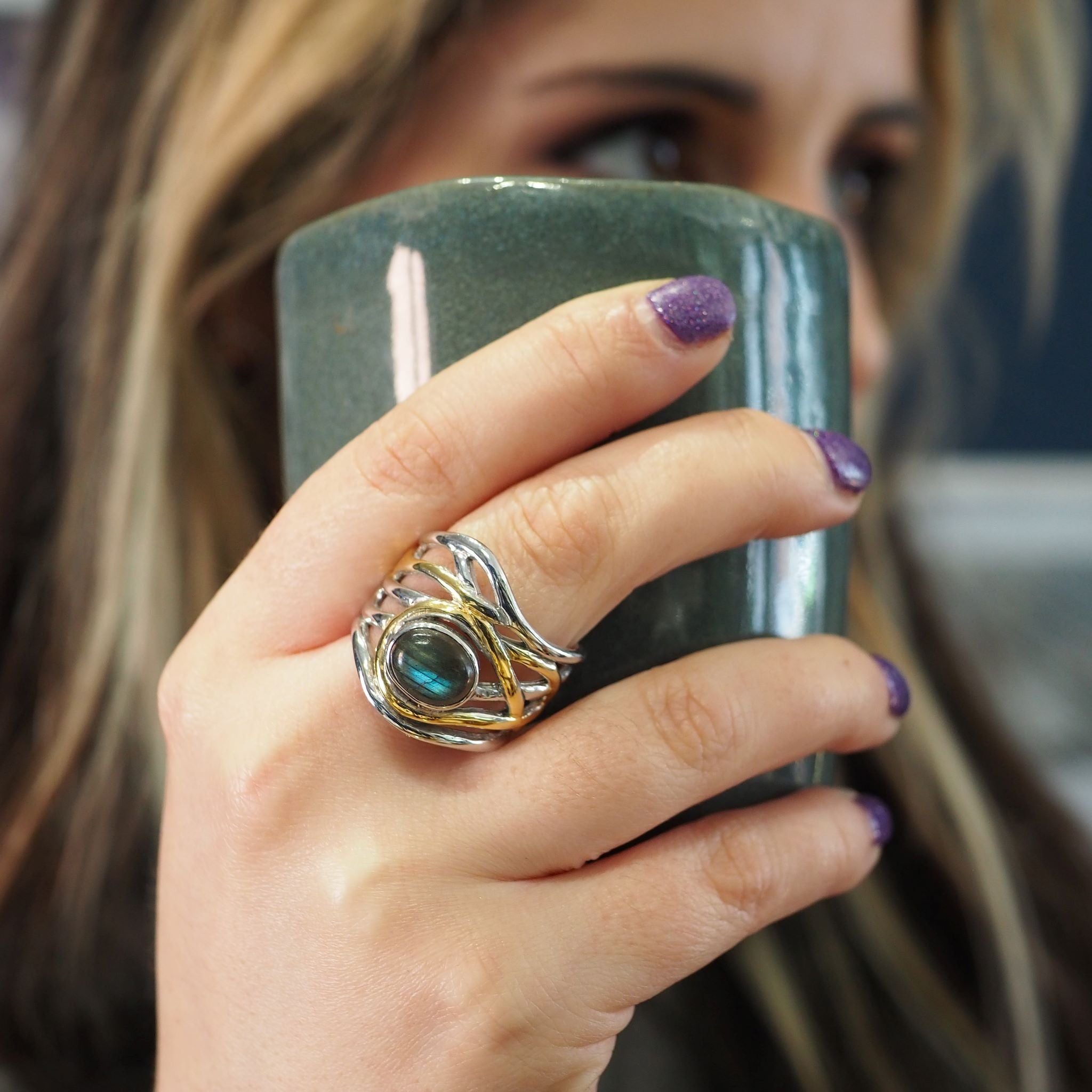 Close-up of a person holding a green ceramic mug with their right hand. They are wearing a large, intricate silver and gold chunky statement ring featuring a blue-green stone from the **Mayhem Ring in various gemstones by Gallardo & Blaine Designs**. The 