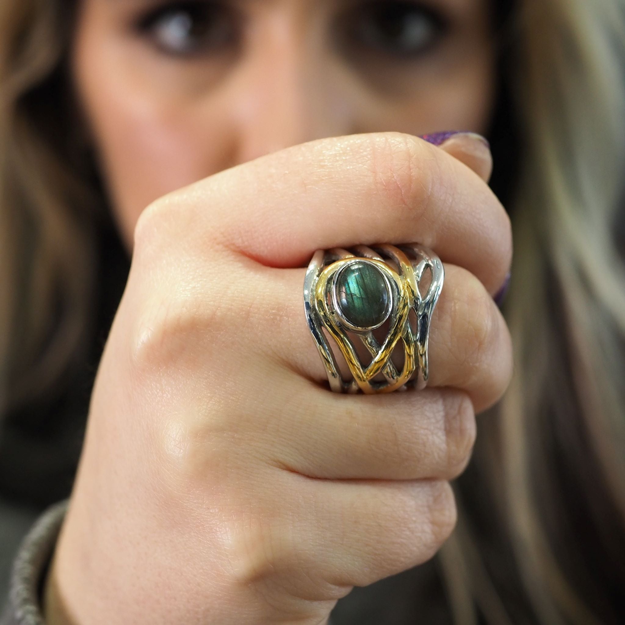 A close-up image of a person showing their hand with a striking, medieval-inspired chunky statement ring. The Mayhem Ring in various gemstones by Gallardo & Blaine Designs has an ornate design with intertwined silver and gold bands, featuring a large, rou