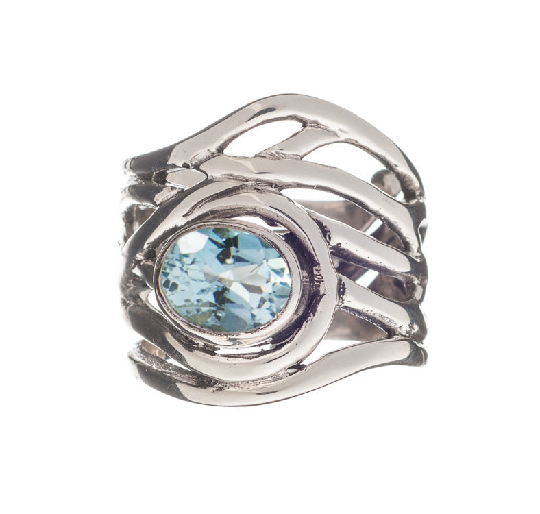 The Mayhem Ring in various gemstones from Gallardo & Blaine Designs features an intricate, wavy design and a large, round blue gemstone set in the center. The stone is faceted, catching the light and adding sparkle to this chunky statement ring reminiscen