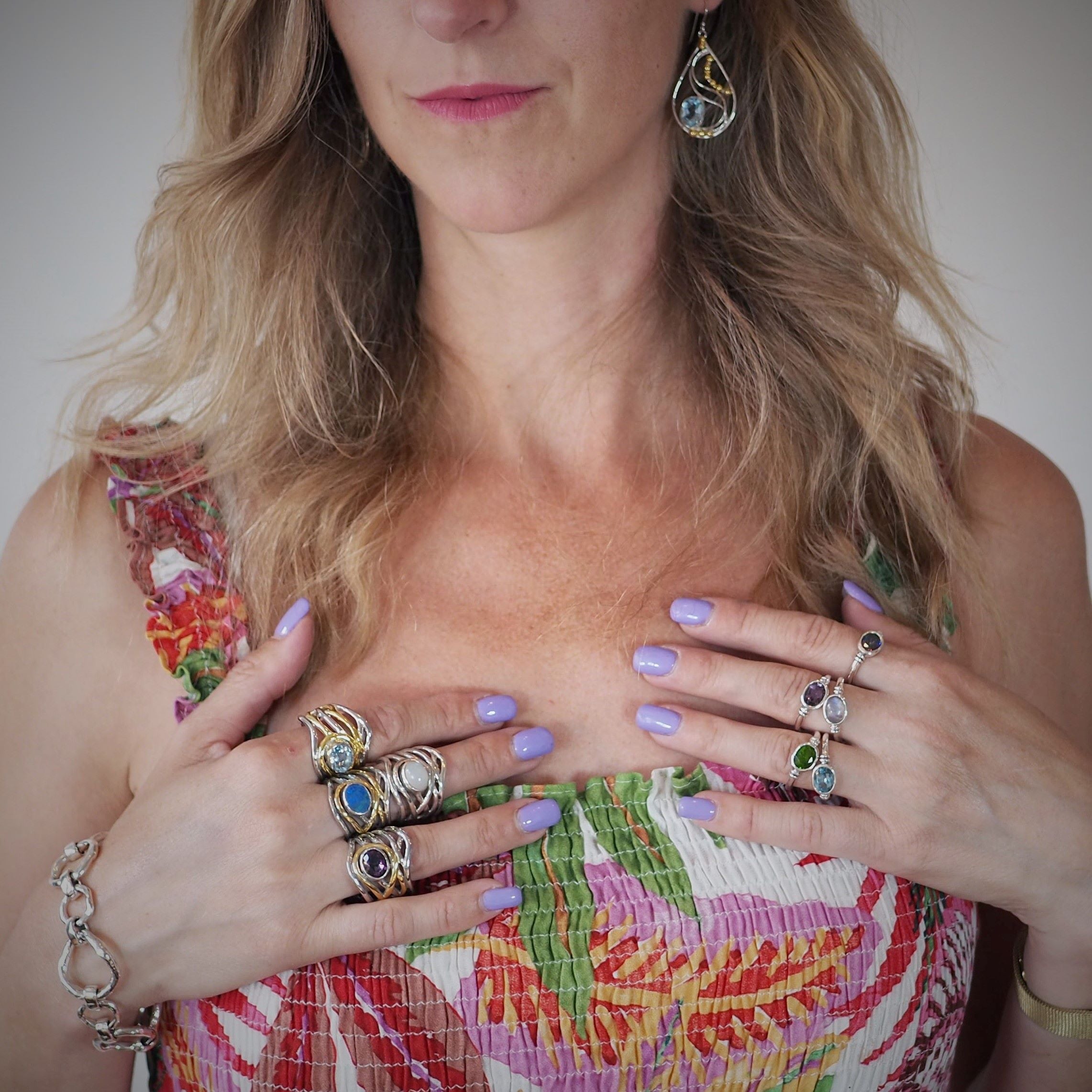 A woman with lavender-painted nails wears multiple Gallardo & Blaine Designs' Mayhem Rings in various gemstones. She has long, wavy blonde hair and dons a colorful, patterned top with thin straps. She also wears a bracelet and dangling earrings that match