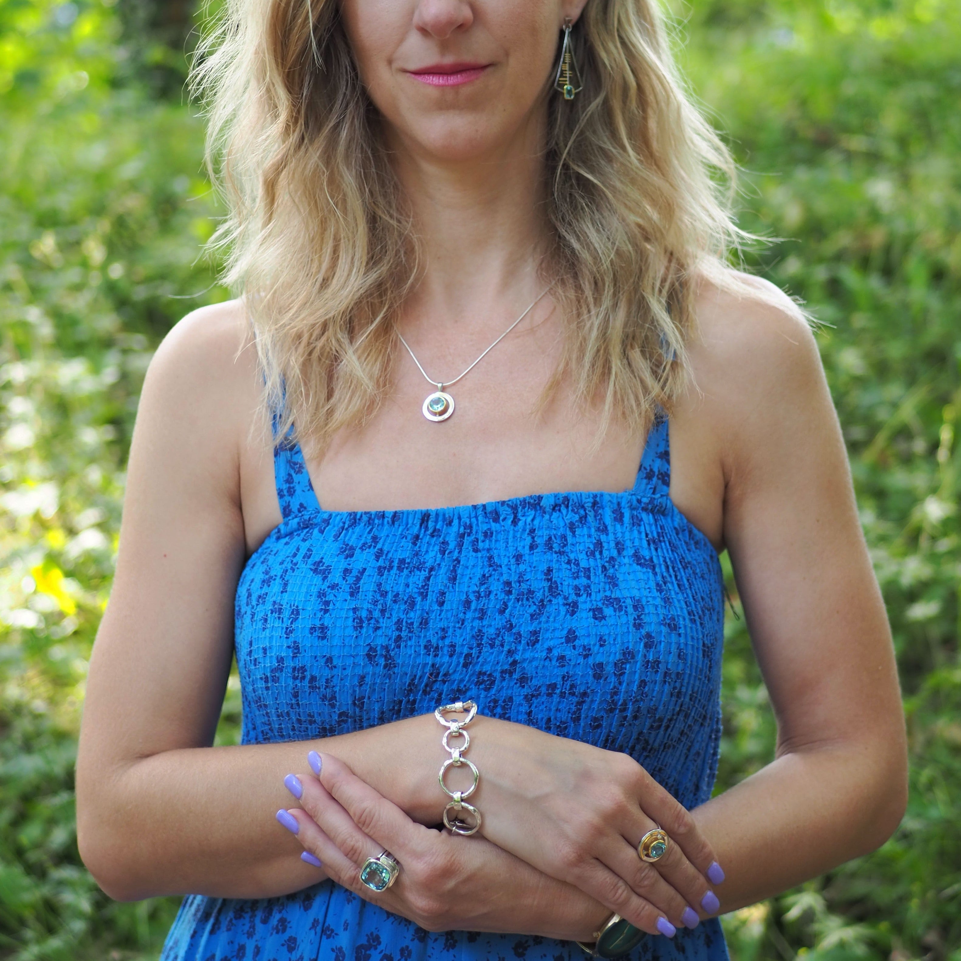 A woman with wavy blonde hair stands outdoors wearing a blue patterned sundress. She has a necklace, earrings, and a statement Gallardo & Blaine Designs Sea Honey Bracelet Silver with large, thick silver links. Her nails are painted lilac, and she has mul
