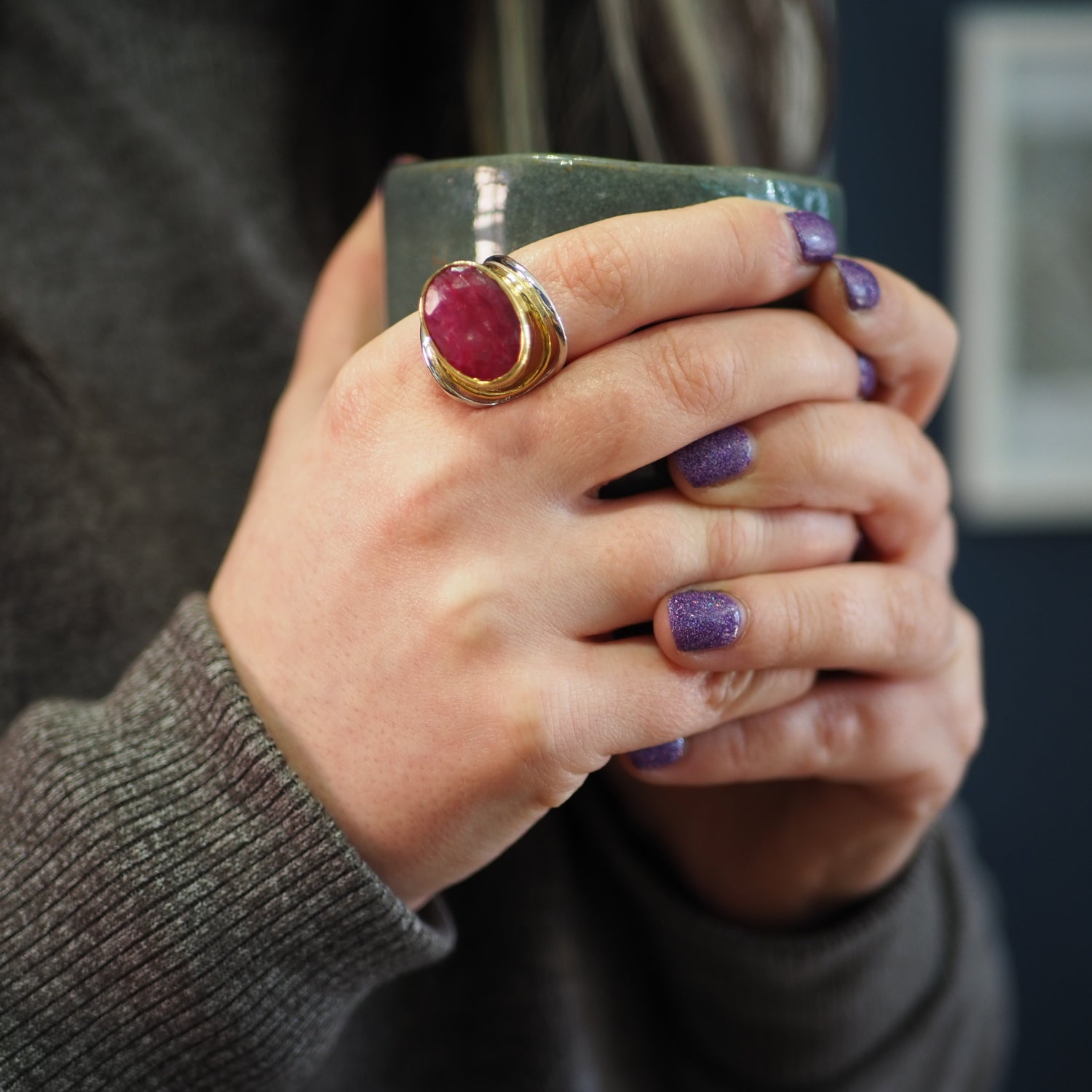 A person with purple glittery nails holds a green mug, wearing Gallardo & Blaine Designs' Party Ring with a red gemstone on an adjustable band, and is dressed in a gray sweater.