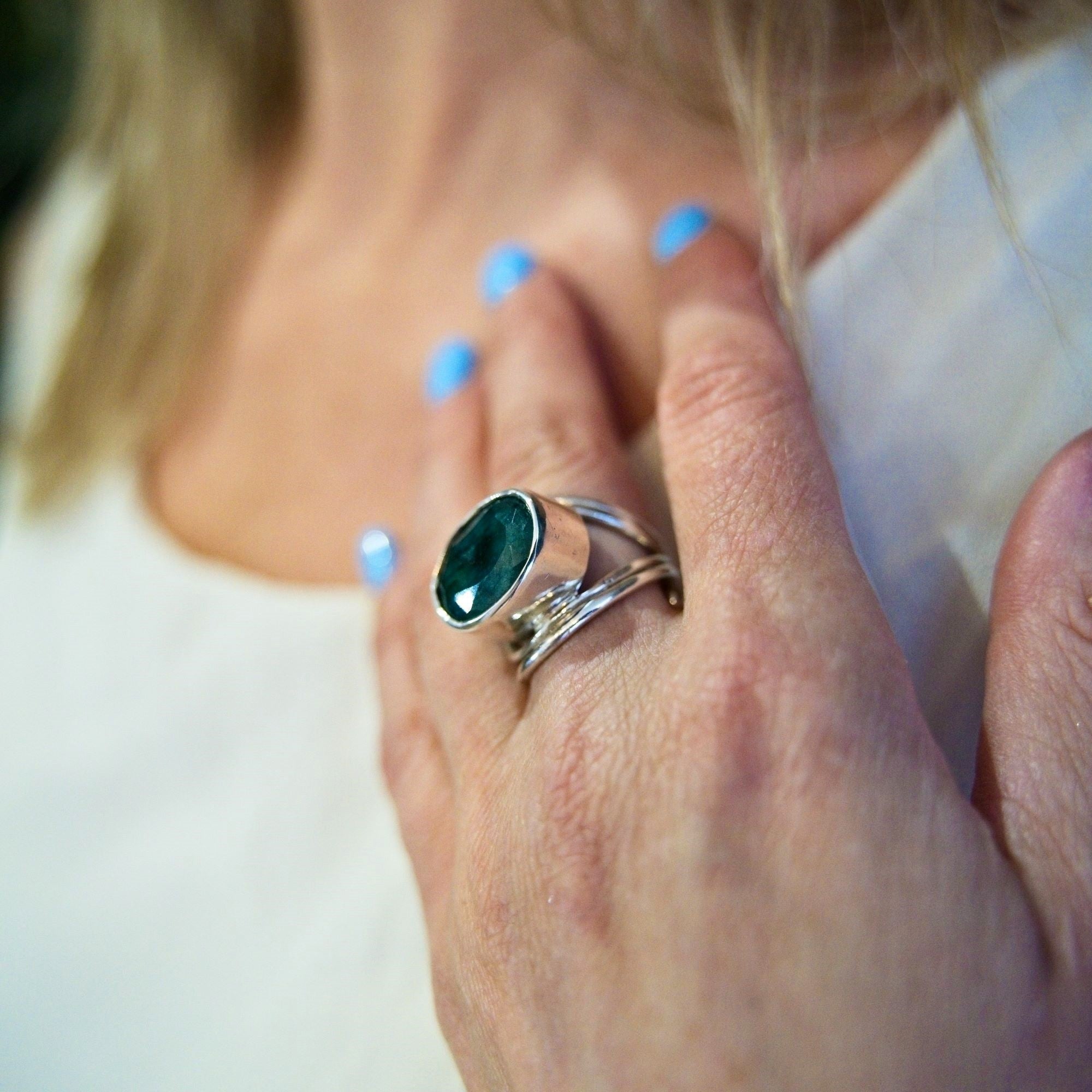 Close-up of a hand with light blue nail polish showcasing Gallardo & Blaine Designs' Party Ring—a chunky silver statement piece featuring a dark green gemstone. This adjustable accessory rests gracefully on a shoulder against white fabric.