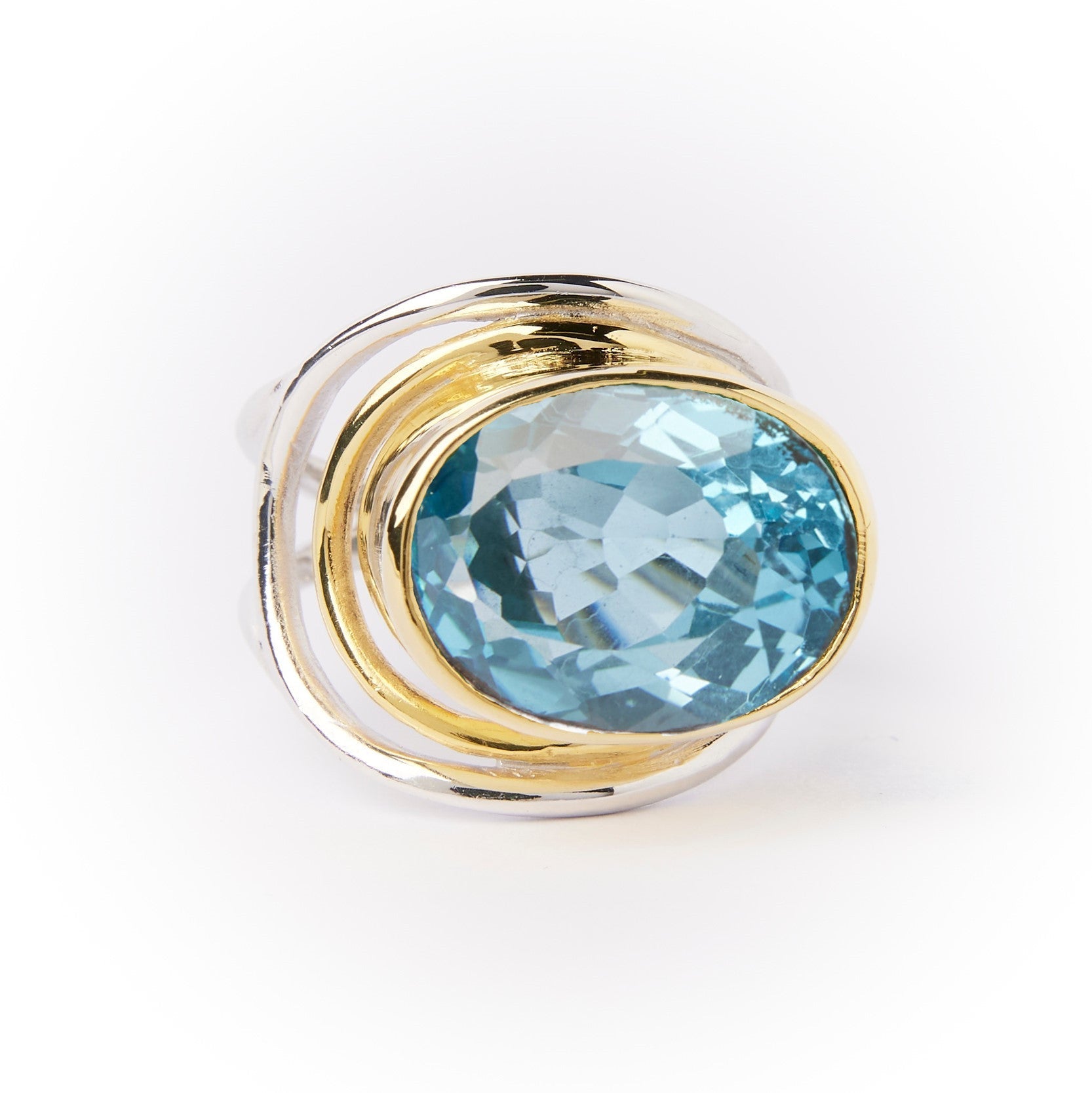A **Party Ring in various gemstones** featuring a large, round, faceted blue statement gemstone in the center by **Gallardo & Blaine Designs**. The gemstone is encircled by swirling, overlapping gold bands, enhancing the ring's elegant and sophisticated d
