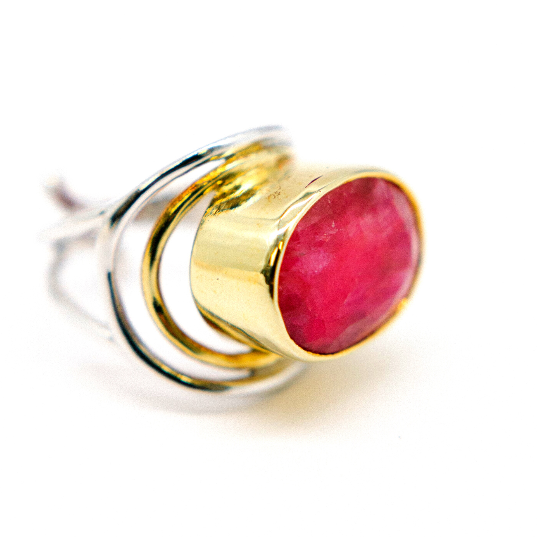 A close-up of Gallardo & Blaine Designs' Party Ring showcasing a chunky statement with a large oval pink gemstone in a gold bezel. The band features intertwining silver loops, offering a unique and elegant design against a plain white background.