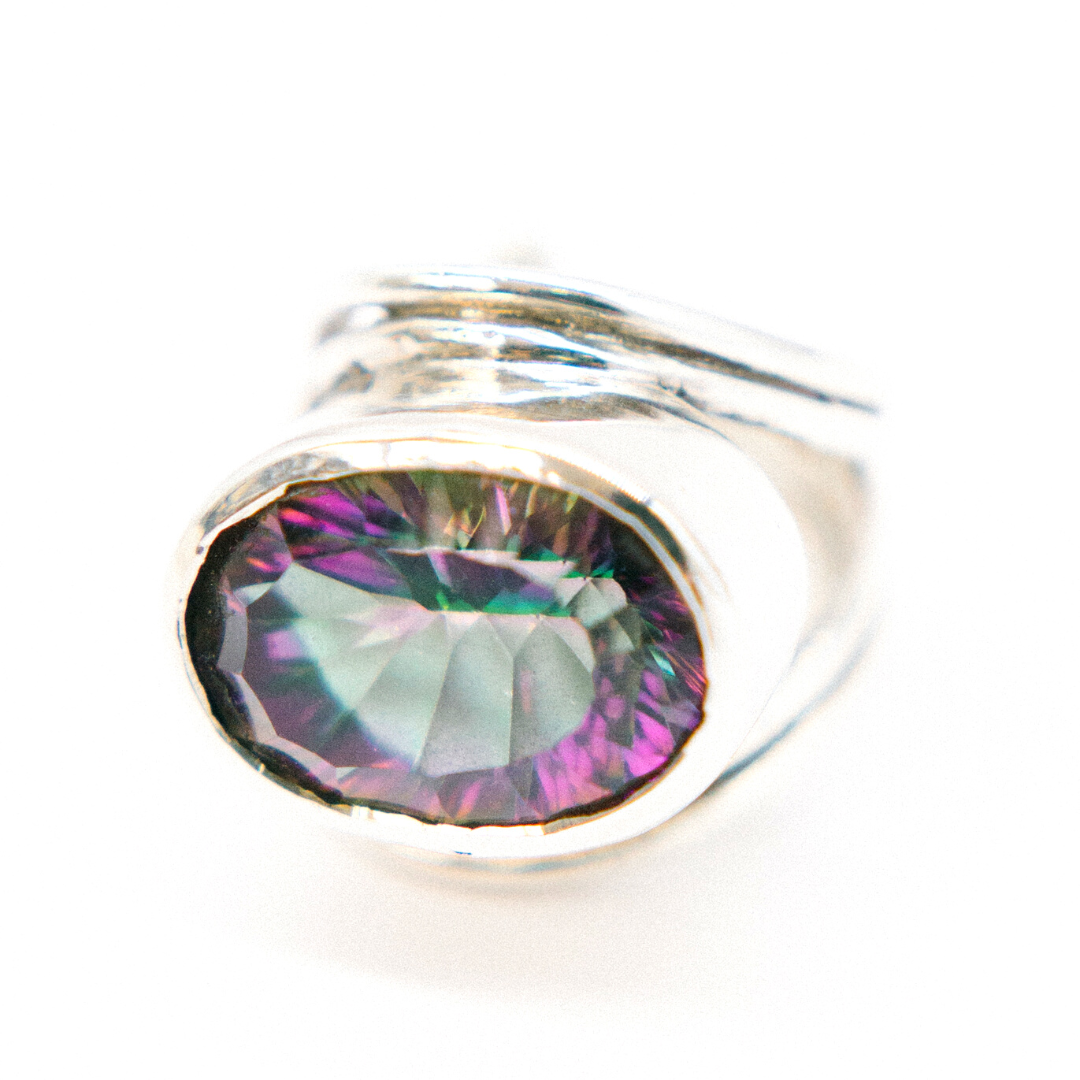 A close-up image of a statement gemstone ring featuring a large, multicolored gemstone with hues of green, purple, and hints of blue, set in a simple bezel setting. The background is plain white, emphasizing the Gallardo & Blaine Designs Party Ring in var