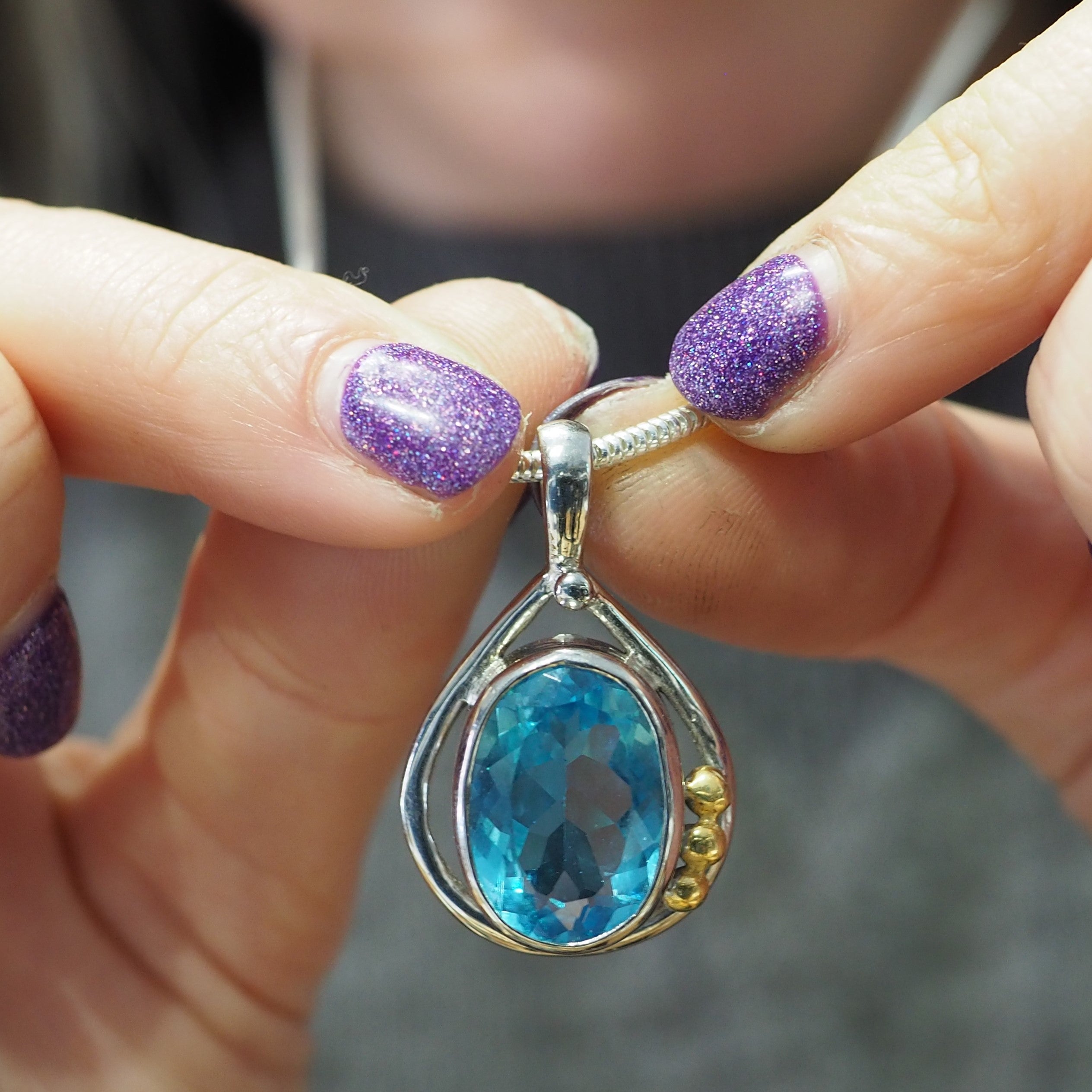 Close-up of hands holding a necklace with a pendant. The Poppy Pendant Small in various gemstones by Gallardo & Blaine Designs features a large, oval-cut blue gemstone set in a sterling silver teardrop-shaped frame adorned with three small gold accents. T