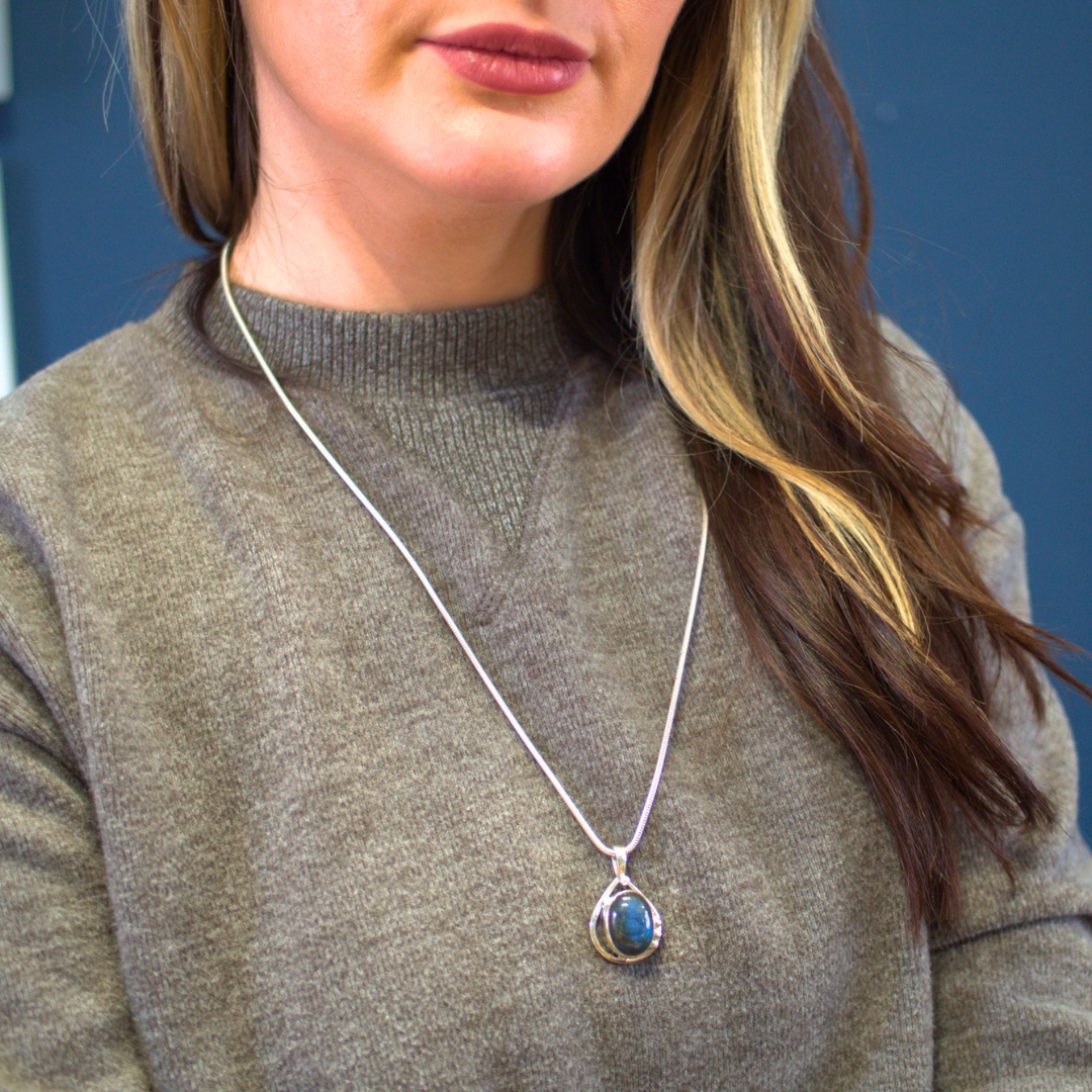 A person wearing a grey sweater is shown from the shoulders up. They have long brown hair with blonde highlights and are wearing a Gallardo & Blaine Designs sterling silver necklace with a large blue Poppy Pendant Small in various gemstones. The backgroun