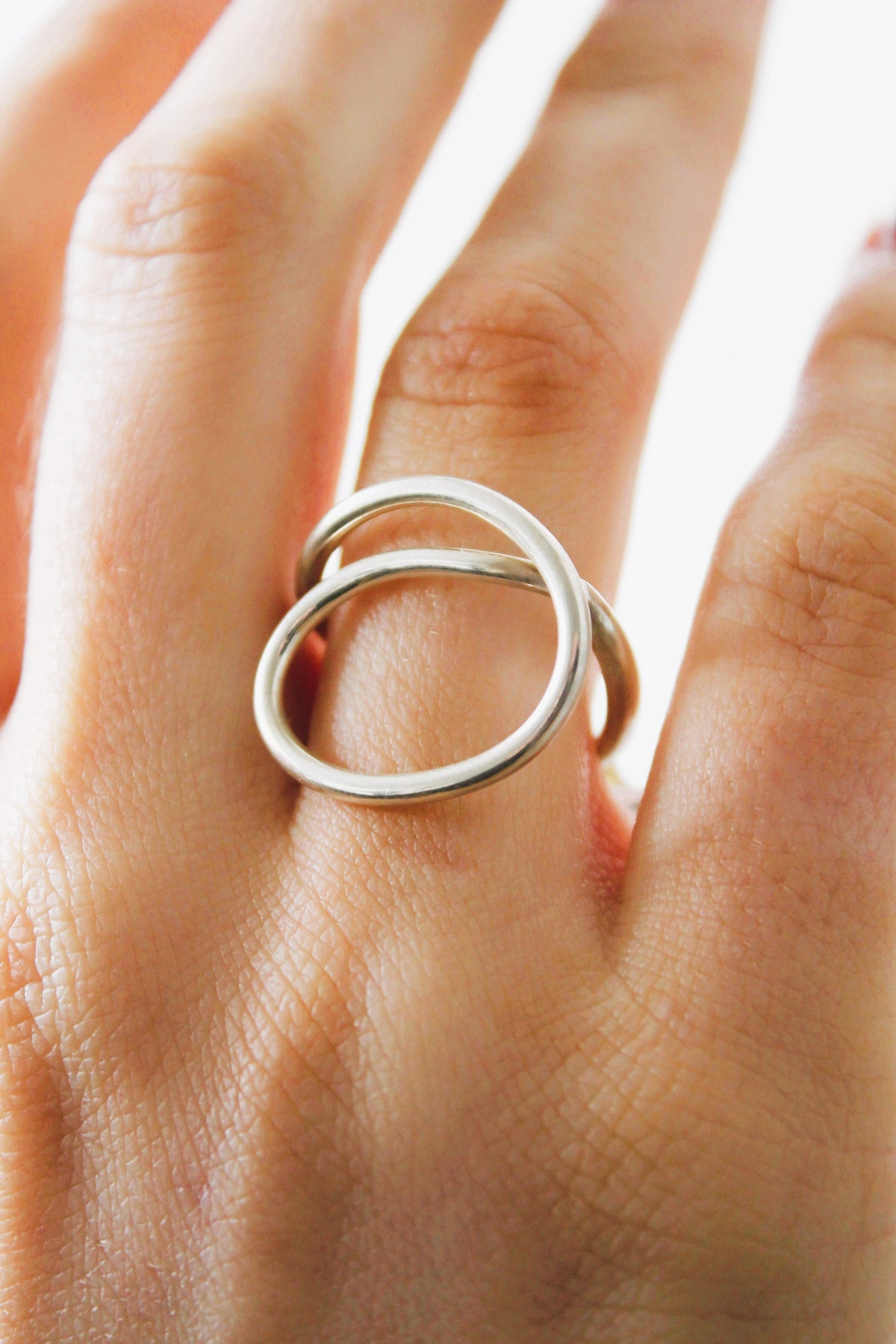 A close-up of a hand wearing the Sea Honey Ring by Gallardo & Blaine Designs, a minimalist sterling silver piece with two interlocking circles, shown against a soft light background.
