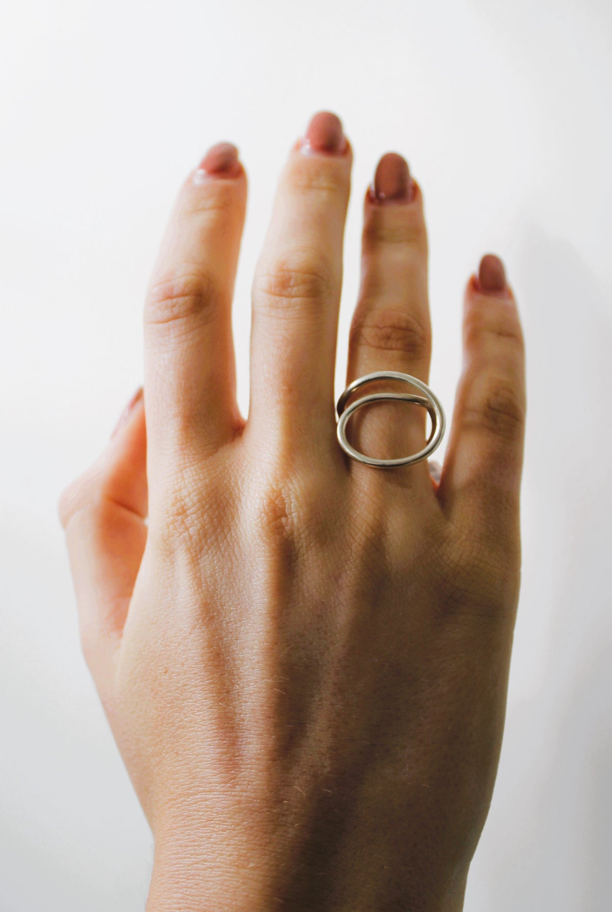 A hand with neatly manicured nails displays the Gallardo & Blaine Designs Sea Honey Ring, a minimalist sterling silver piece with two overlapping ovals, on the ring finger against a plain white background.