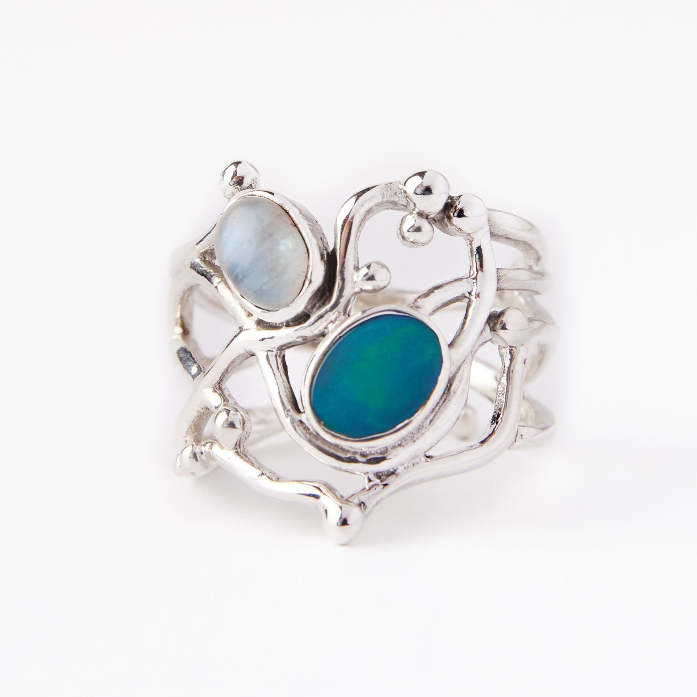 The Seaweed Ring in various gemstones by Gallardo & Blaine Designs in sterling silver features an abstract, wavy design with two oval stones. One stone is a pale, milky color while the other is a vibrant, deep blue-green. The ring's design includes organi