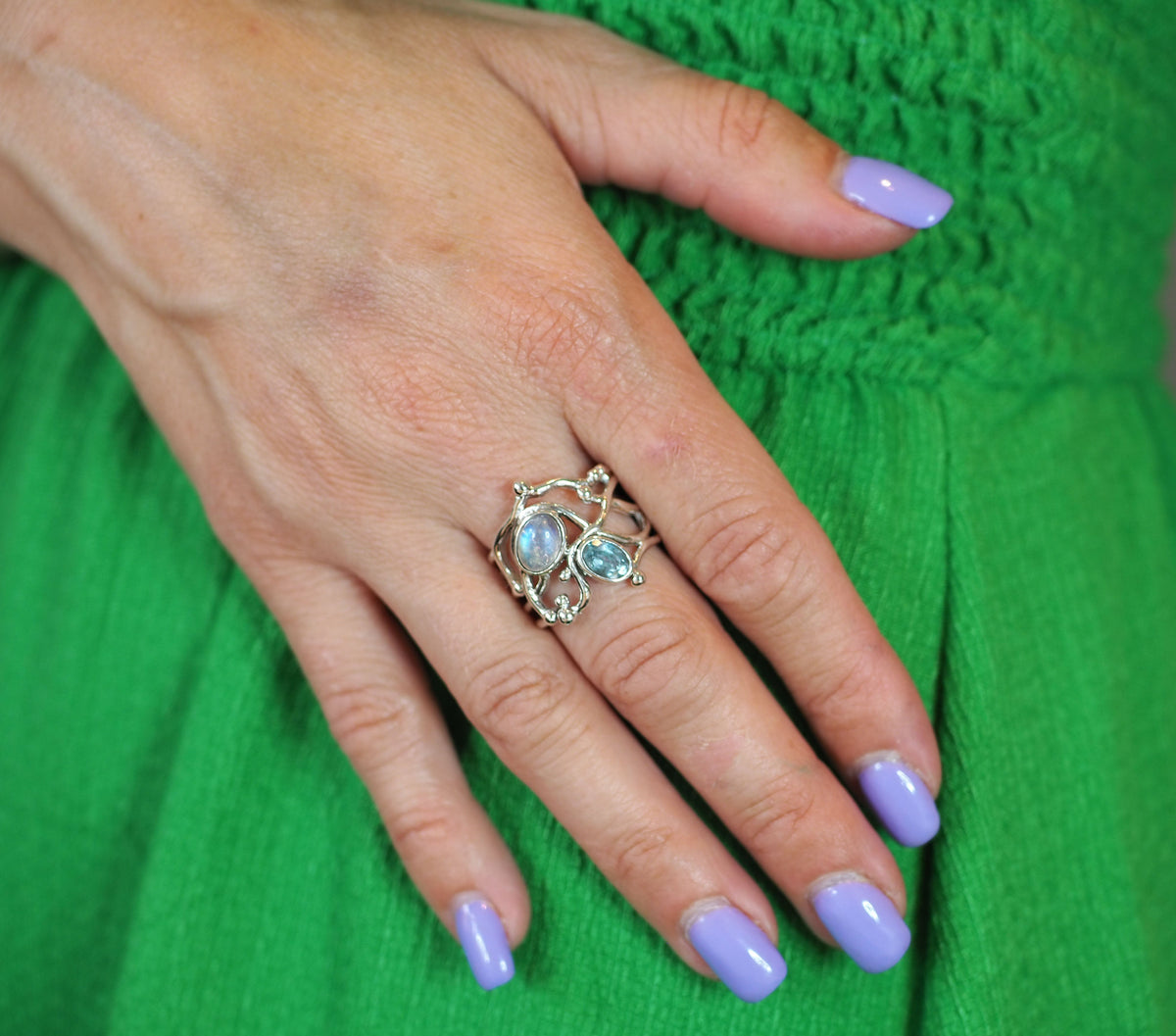 A hand with light purple nails rests on bright green fabric, showcasing Gallardo & Blaine Designs' Seaweed Ring in gold vermeil with two light blue gemstones and an intricate design.