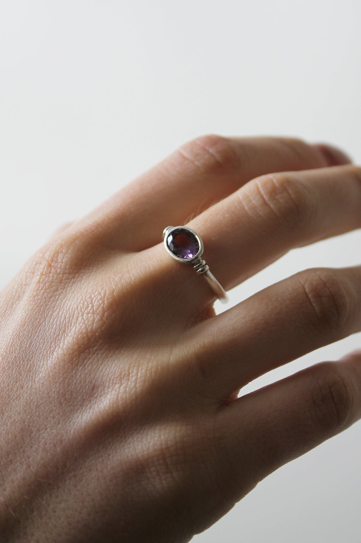 The Senna Ring by Gallardo & Blaine Designs, featuring a small round purple gemstone, is worn on the left ring finger against a plain light background.
