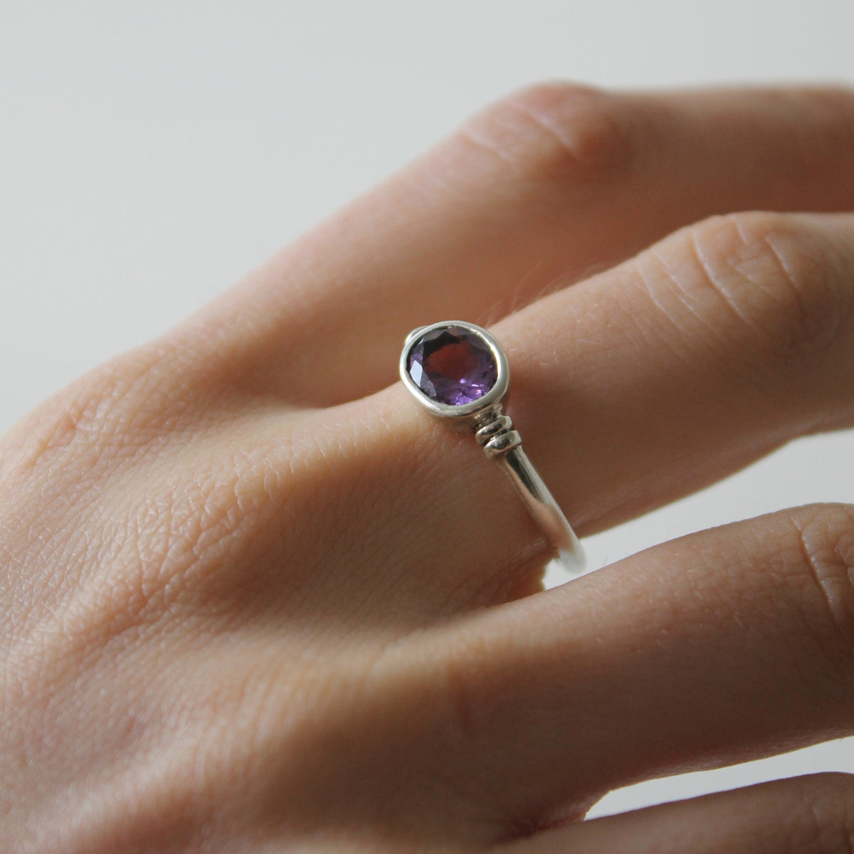 A close-up of a hand wearing the Gallardo & Blaine Designs Senna Ring, shown here in silver with a round purple gemstone in a simple bezel setting.