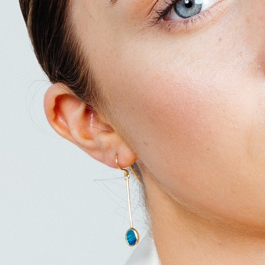 Close-up of a person’s face showing a blue eye, part of their cheek, and ear. They are wearing Sequola Earrings in various gemstones from Gallardo & Blaine Designs: long, slender sterling silver dangle earrings with a blue circular gem. The person's skin 