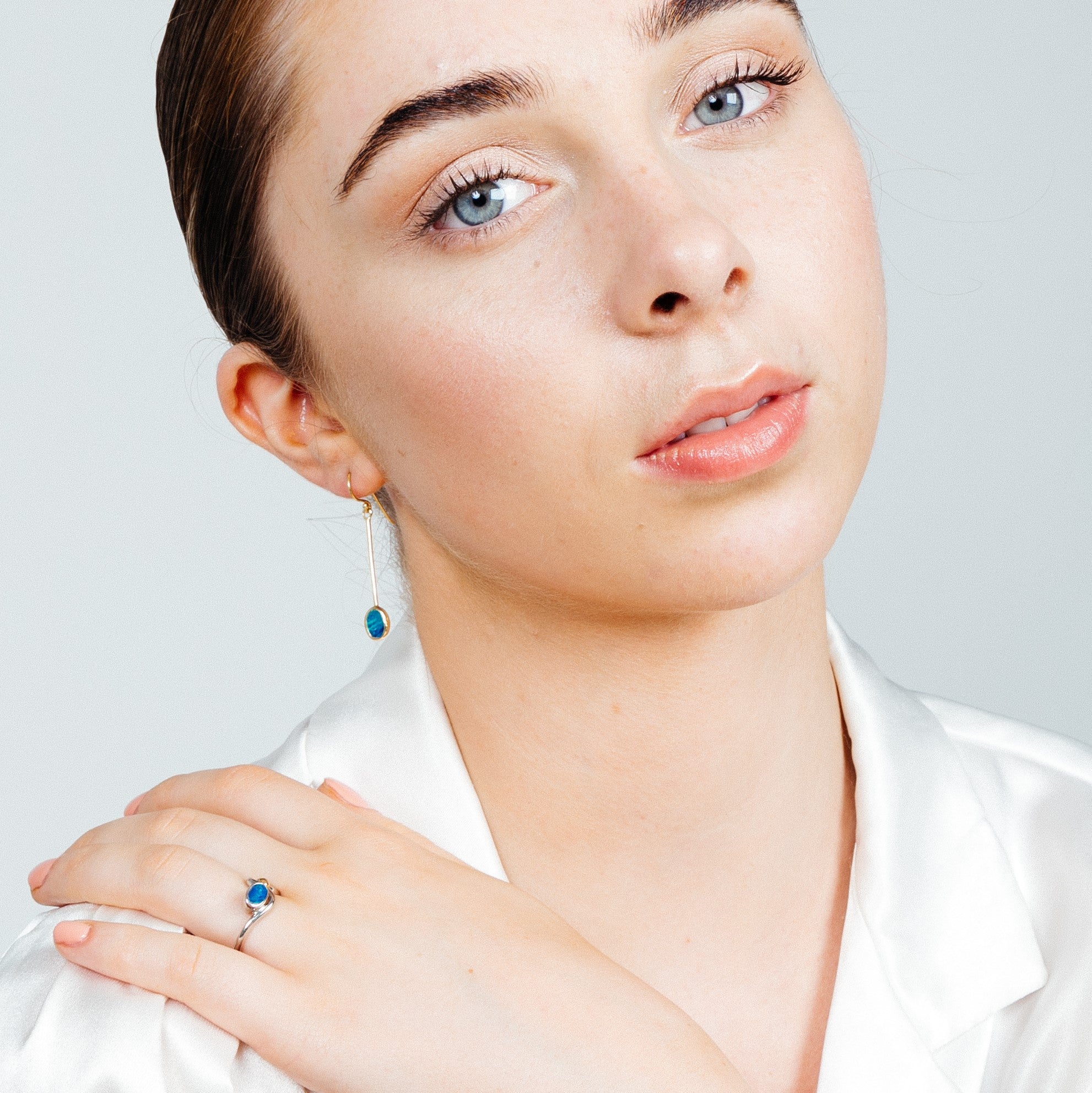 A person with light skin, light blue eyes, and dark hair pulled back is wearing a white blouse. They are adorned with Sequola Earrings in various gemstones by Gallardo & Blaine Designs and a ring, both featuring blue gemstones. The person gazes softly at 