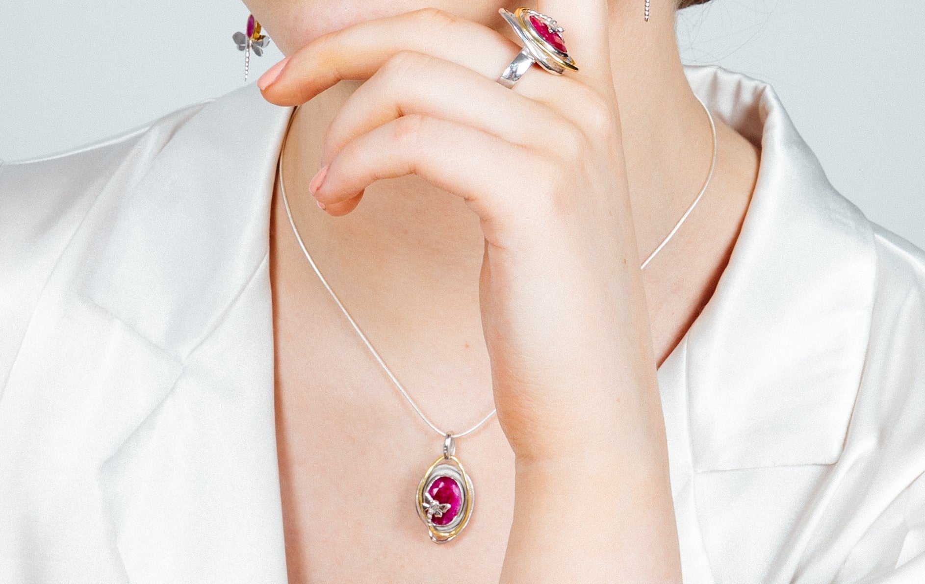 A person wearing a white shirt is adorned with jewelry, including a silver ring with a red gemstone on their middle finger, a Lagoon Pendant Small in various gemstones by Gallardo & Blaine Designs featuring a matching red gemstone, and silver earrings. Th