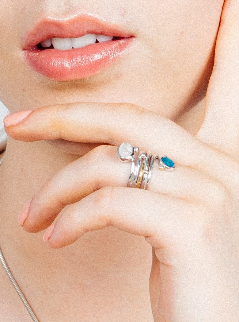 Close-up of a person's lips and hand. The person is wearing four silver rings on their index finger, one with a white stone and another with a unique turquoise design. A Snake Ring in Silver Gold & various gemstones by Gallardo & Blaine Designs graces the