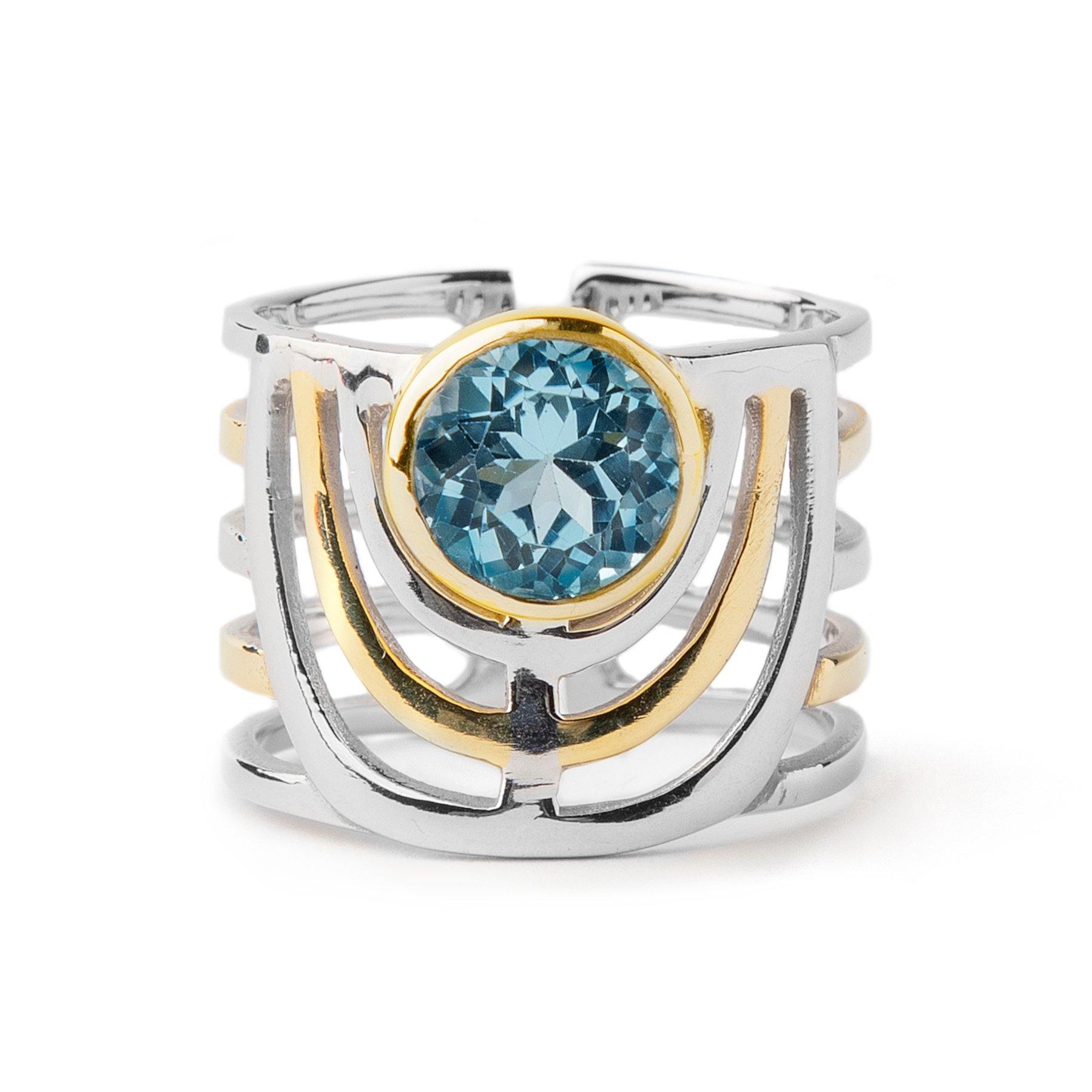 A bold jewellery design, the Solar Ring in various gemstones by Gallardo & Blaine Designs showcases a geometric pattern with a prominent blue gemstone at its heart. The open band structure is accented with gold, artistically framing the gemstone that's en