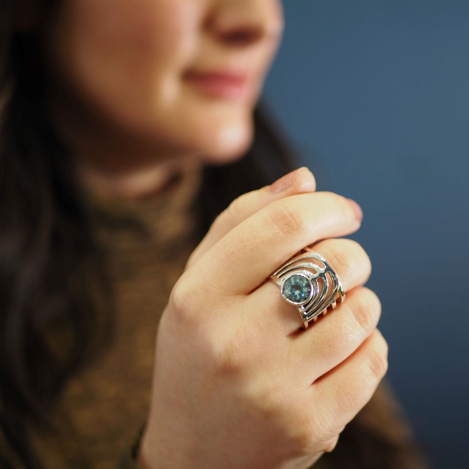 A person holding their hand near their face, wearing a bold jewellery design featuring the Gallardo & Blaine Designs Solar Ring in various gemstones with multiple bands and a round blue gemstone. The background is blurred, making the chunky gemstone ring 
