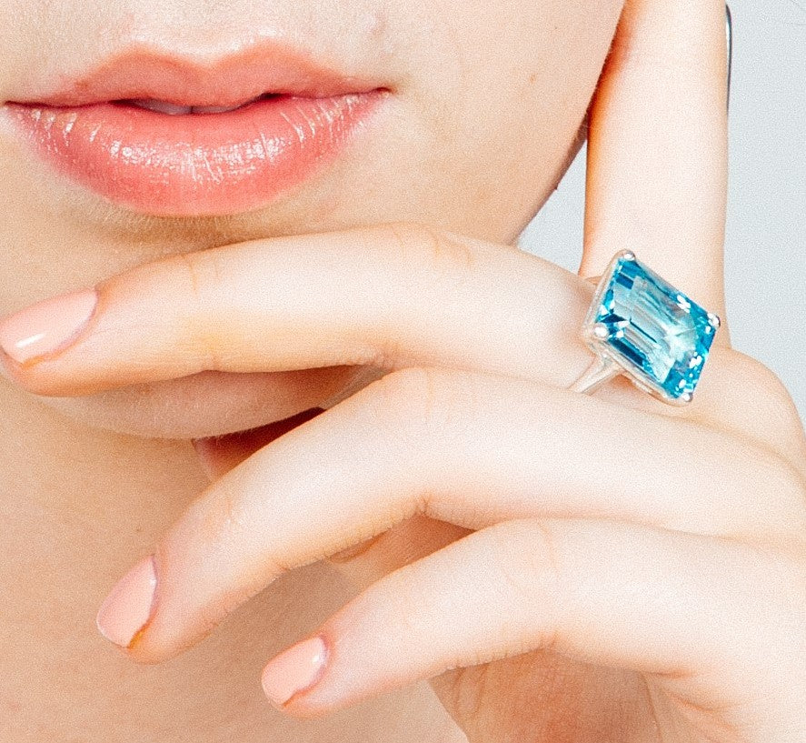 Close-up of a woman's lips and fingers. She is wearing a large, rectangular-cut Stellar Ring in Hydro Blue Topaz by Gallardo & Blaine Designs on her index finger crafted from sterling silver. The nails are manicured with a pale pink color. The background 