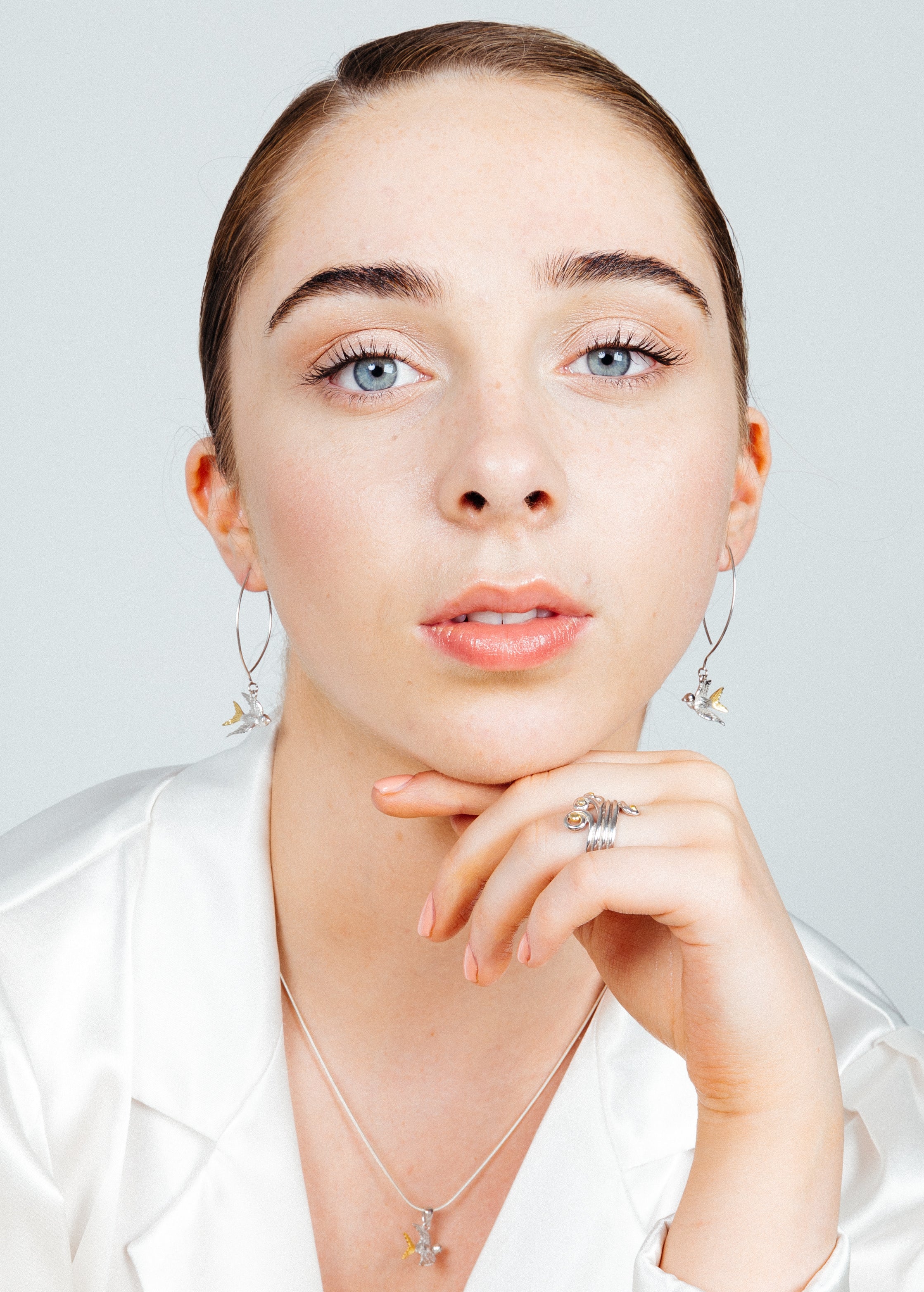 A person with light skin, light blue eyes, and brown hair, pulled back neatly, is wearing silver hoop earrings with small dangling charms, a sterling silver snake chain necklace with a Wildlife Pendant - Swallow With Chain by Gallardo & Blaine Designs, an