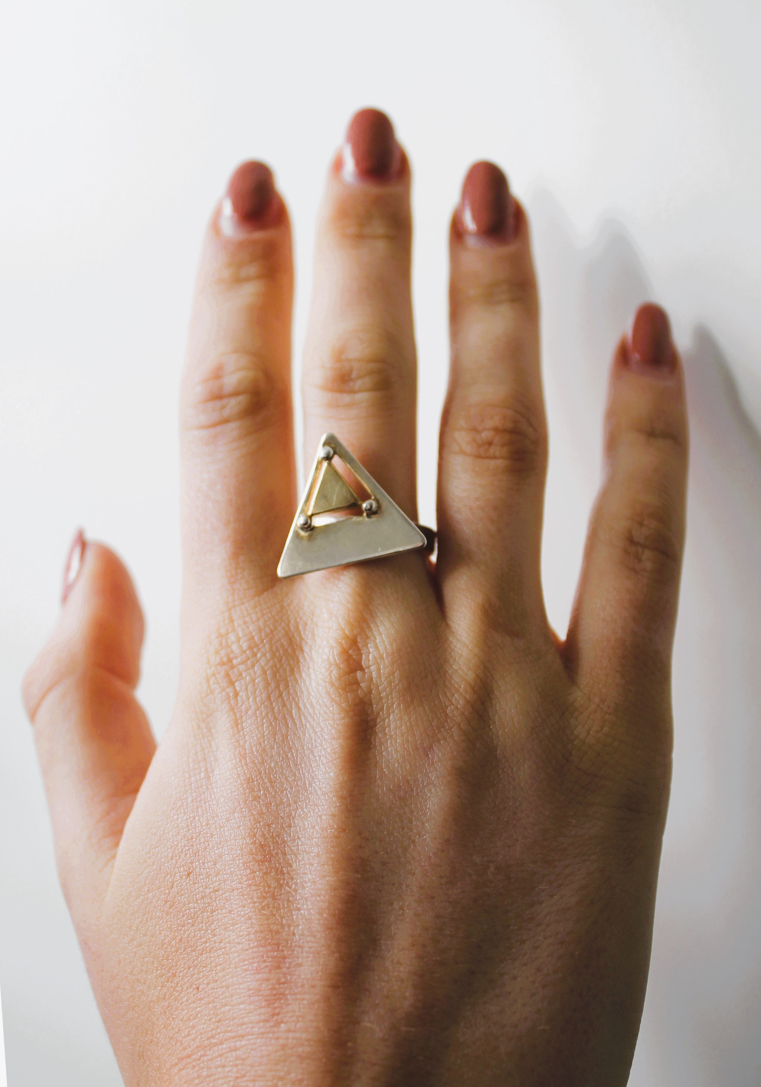 A hand with neatly manicured nails wears the Triune Ring by Gallardo & Blaine Designs—a sterling silver ring featuring a geometric triangle design—on the middle finger, set against a plain white background.