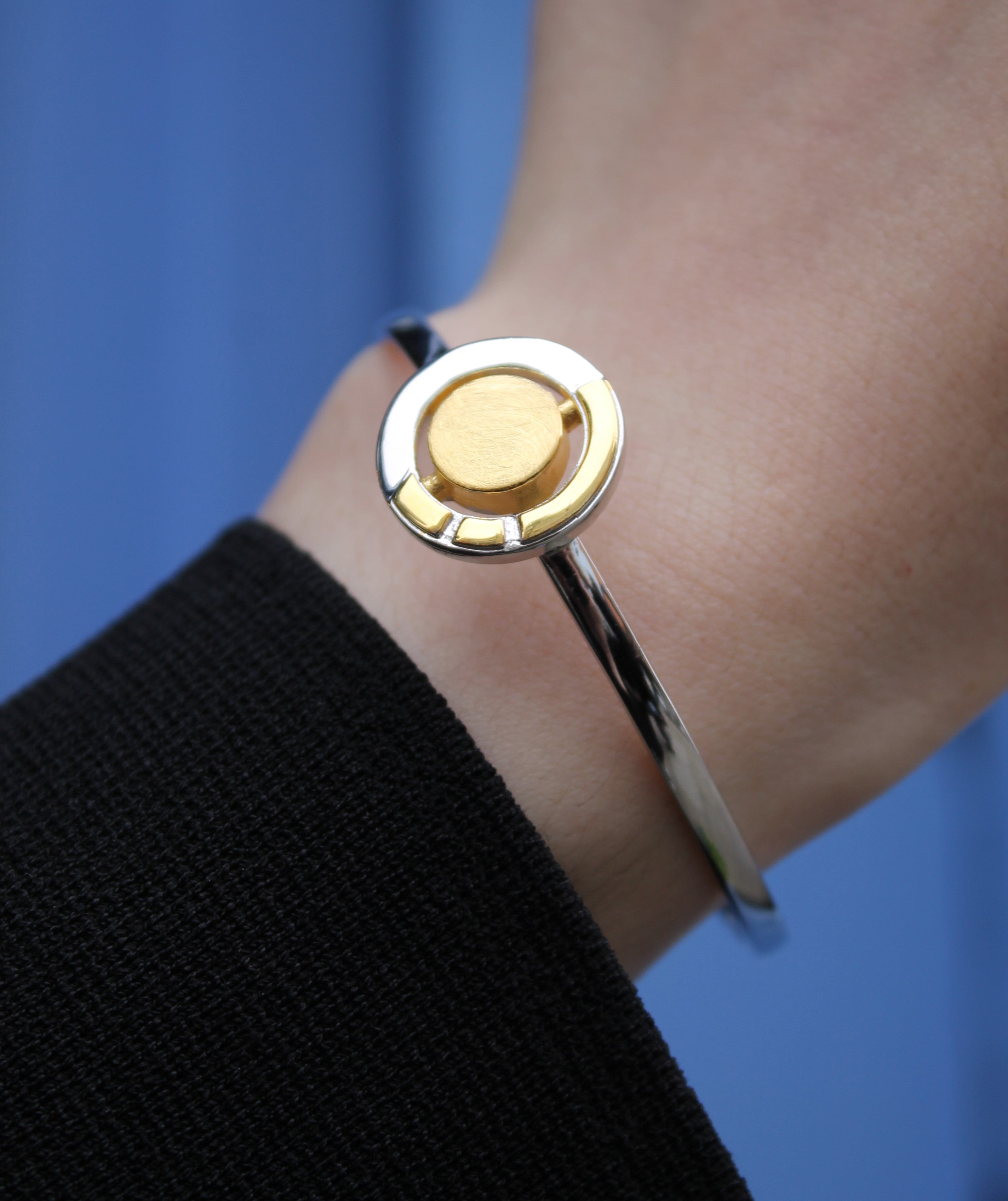 A close-up shows a wrist wearing the Urú Cuff Silver & Gold bracelet by Gallardo & Blaine Designs, featuring a sleek sterling silver band with a round gold accent. The person is dressed in black against a solid blue background.