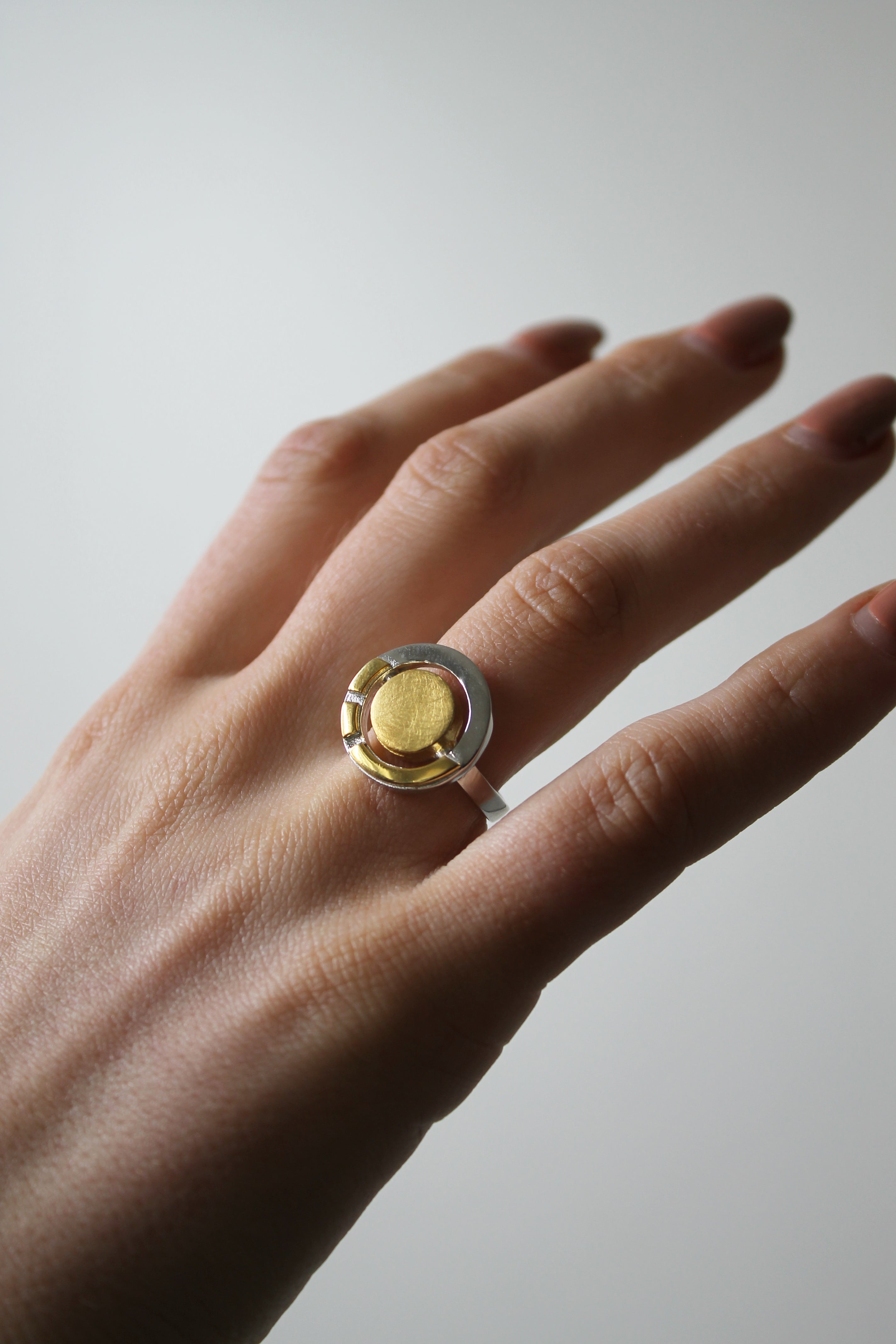 A hand with neatly manicured nails wears the Gallardo & Blaine Designs Urú Ring, a silver Celtic piece with a large, round, gold-colored centerpiece, set against a neutral light background.