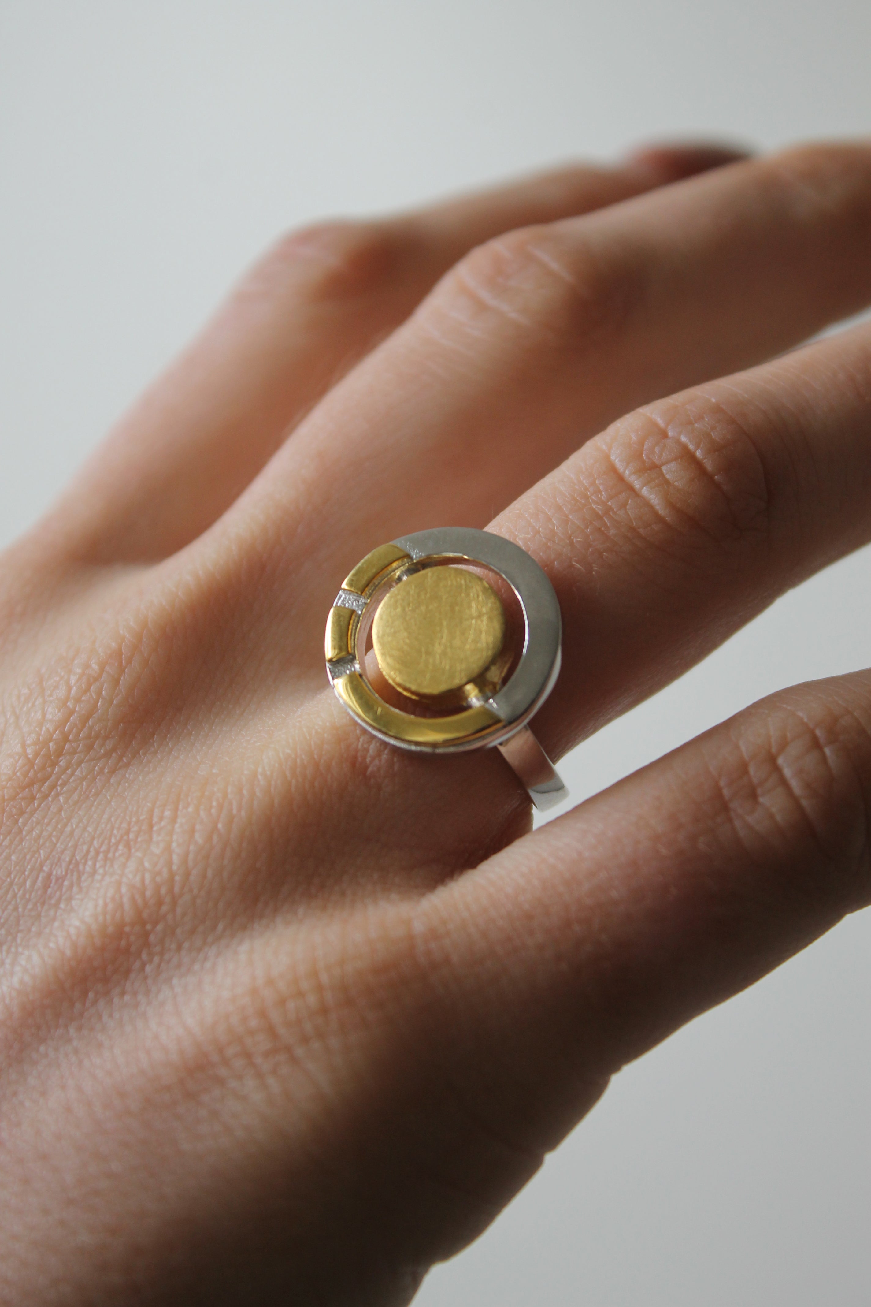 A close-up of a hand wearing the Urú Ring by Gallardo & Blaine Designs, showcasing its central gold disc and mixed metal band. The softly blurred background highlights this striking piece of modern Celtic heritage jewelry.