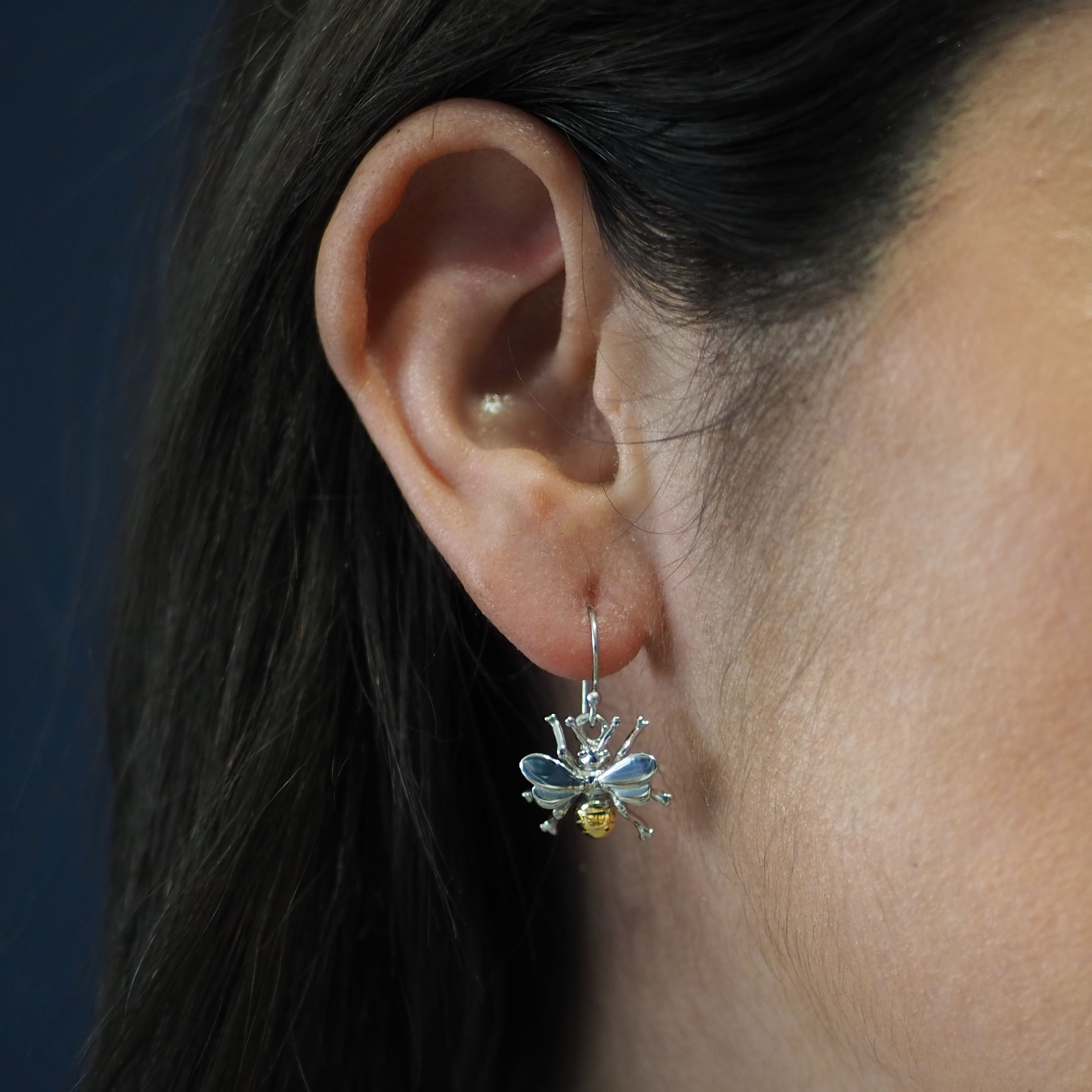 Close-up of a woman's ear wearing Watermelon Tropical's Wildlife Bee Dangle Earring in silver with gold detail, highlighting the metallic finish. The background is dark and blurred.