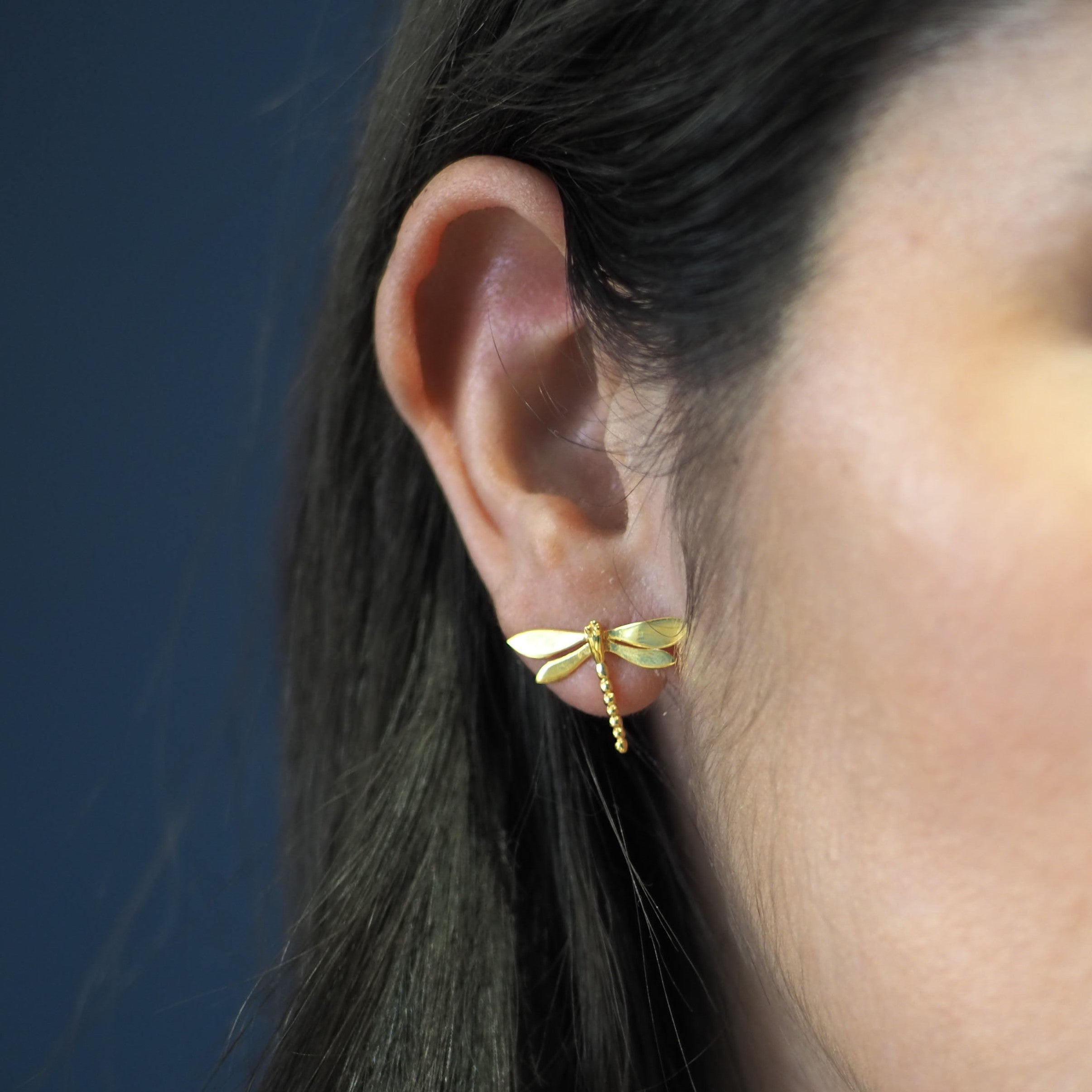 Close-up of a woman wearing Watermelon Tropical's Wildlife Large Dragonfly Studs in gold. Her dark brown hair frames her ear against a blurred, dark blue background, with only part of her face visible. Inspired by nature jewelry.