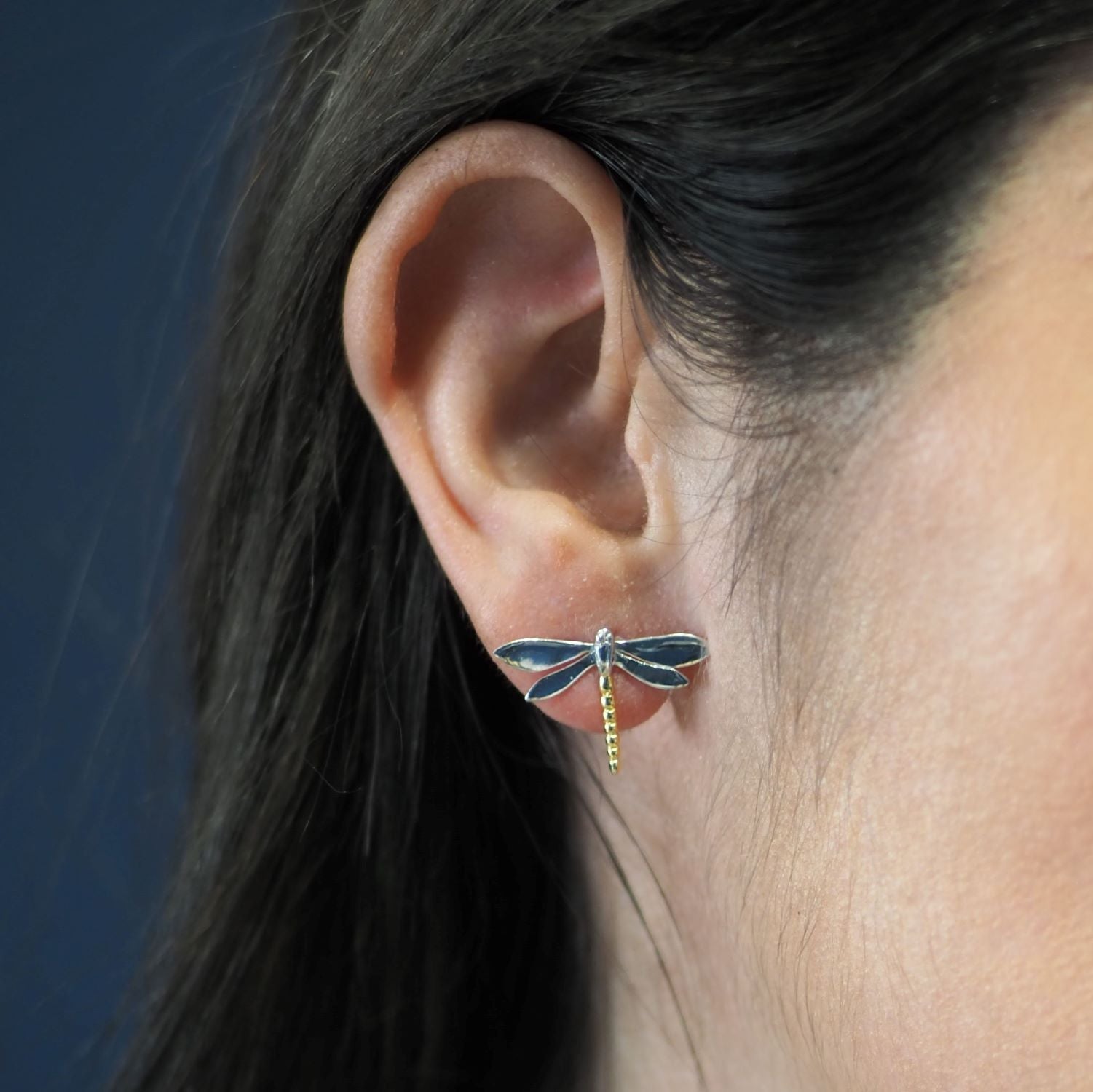 A close-up of a person’s ear wearing Watermelon Tropical Wildlife Large Dragonfly Studs in silver or gold, featuring blue wings and a yellow-striped tail. Both the background and the person’s hair are dark brown.