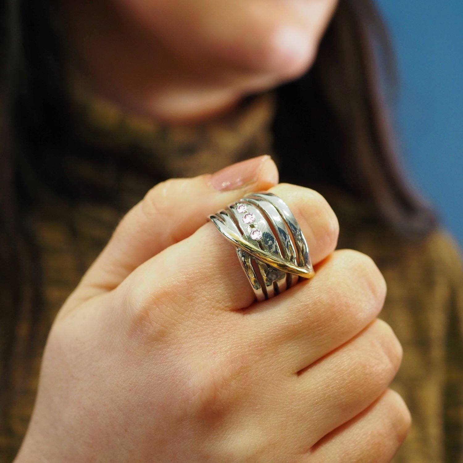 Close-up of a person's hand adorned with Gallardo & Blaine Designs' Adjustable Hidden Gems Ring in Silver & Gold, featuring intertwined bands and sparkling stones. The ring's sterling silver contrasts beautifully with the brown long-sleeve top against a b