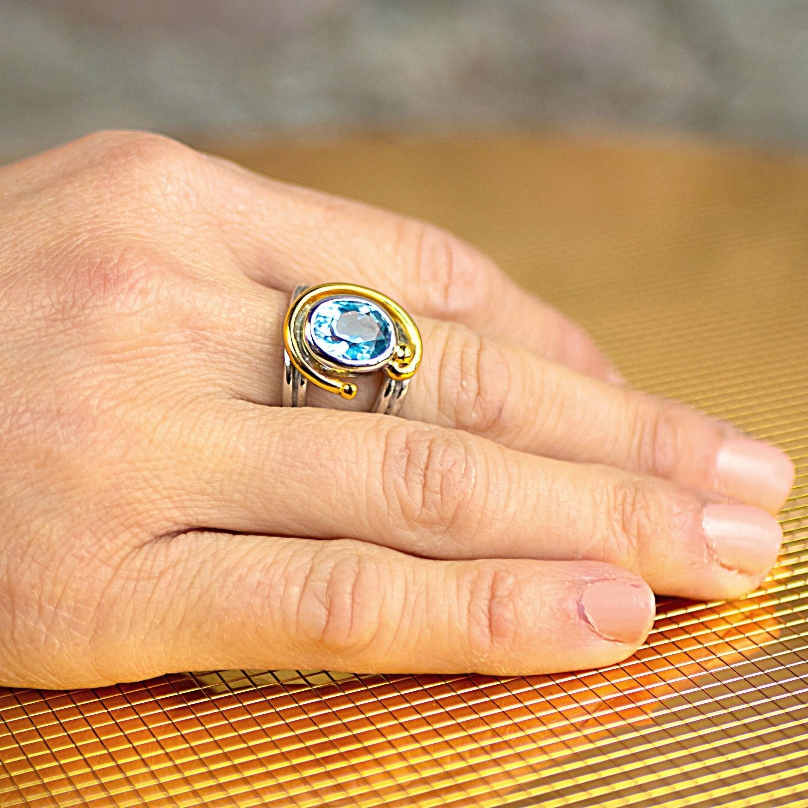 A person's hand with neatly trimmed nails rests on a surface with a grid pattern. The hand is adorned with the Jasmine Ring in various gemstones by Gallardo & Blaine Designs, featuring intricate designs and a large blue gemstone set in the center, complem
