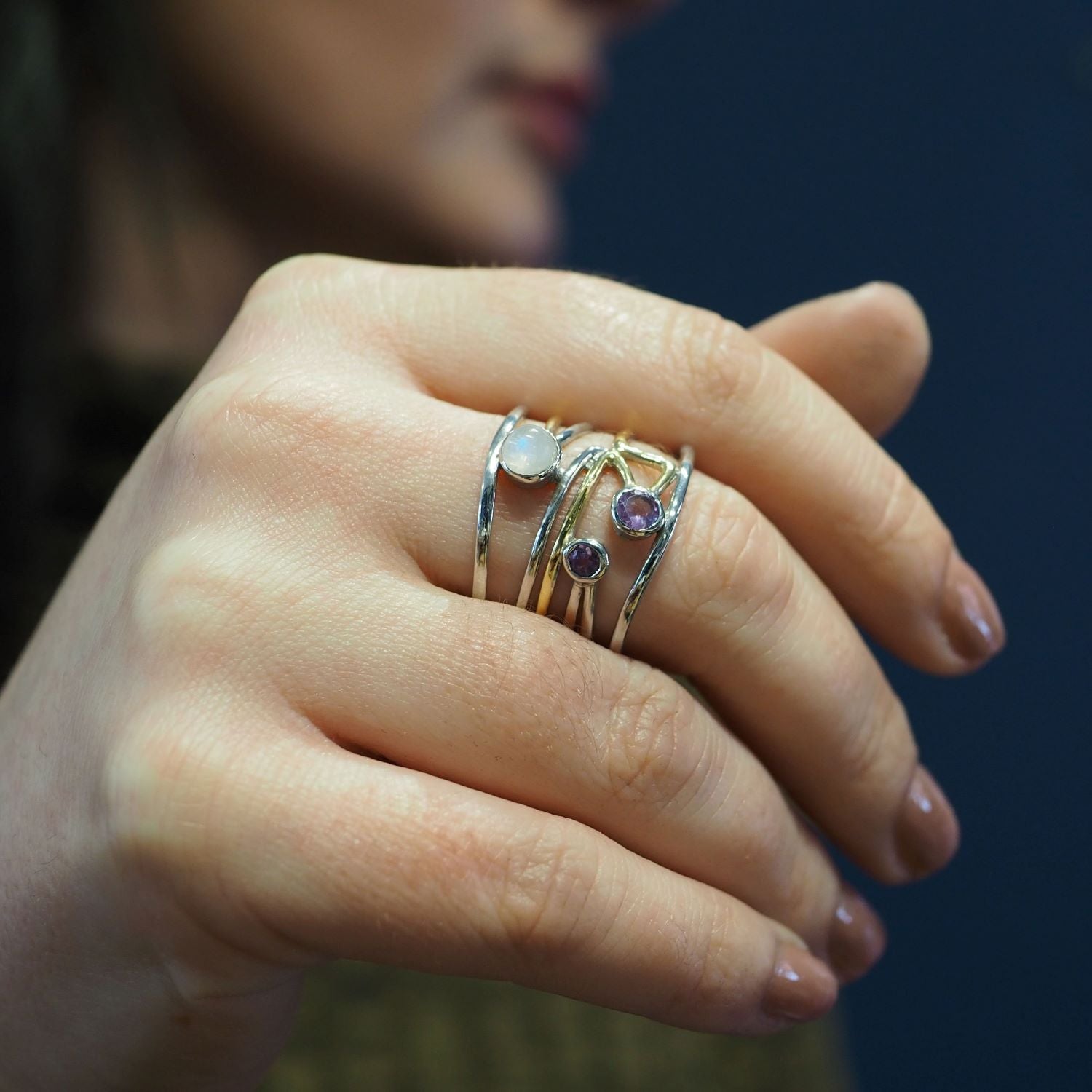 Close-up of a hand adorned with multiple Lattice Rings in various gemstones by Gallardo & Blaine Designs, featuring purple and white stones. The background is blurred, subtly showcasing the person's face and dark hair. The nails are neatly manicured with 