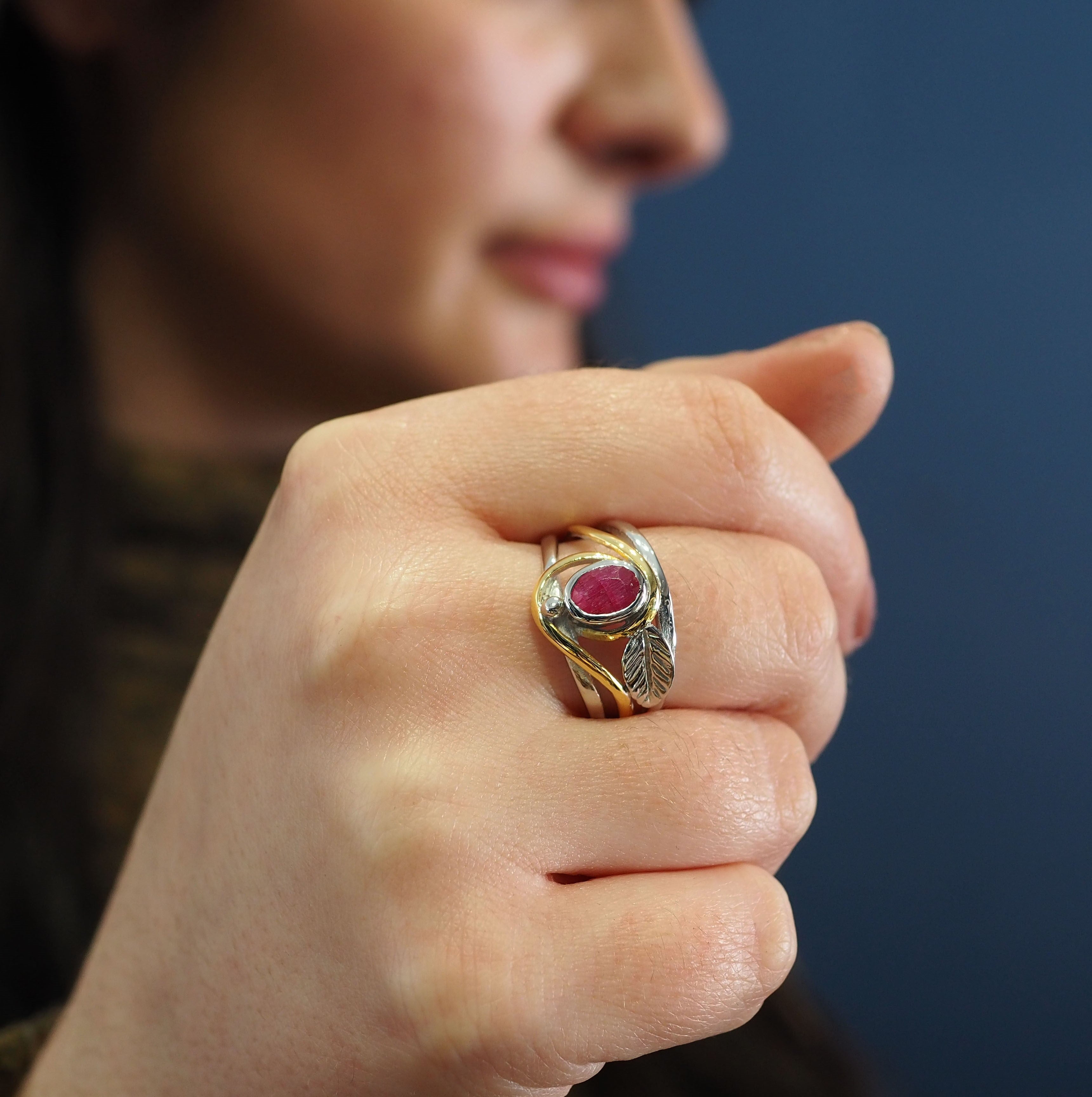 A close-up of a person's hand displaying the Gallardo & Blaine Designs Leaf Ring in Silver Gold & various gemstones, with a red gem and intricate leaf design in silver and gold. The person is partially visible in the background with a blurred face and dar