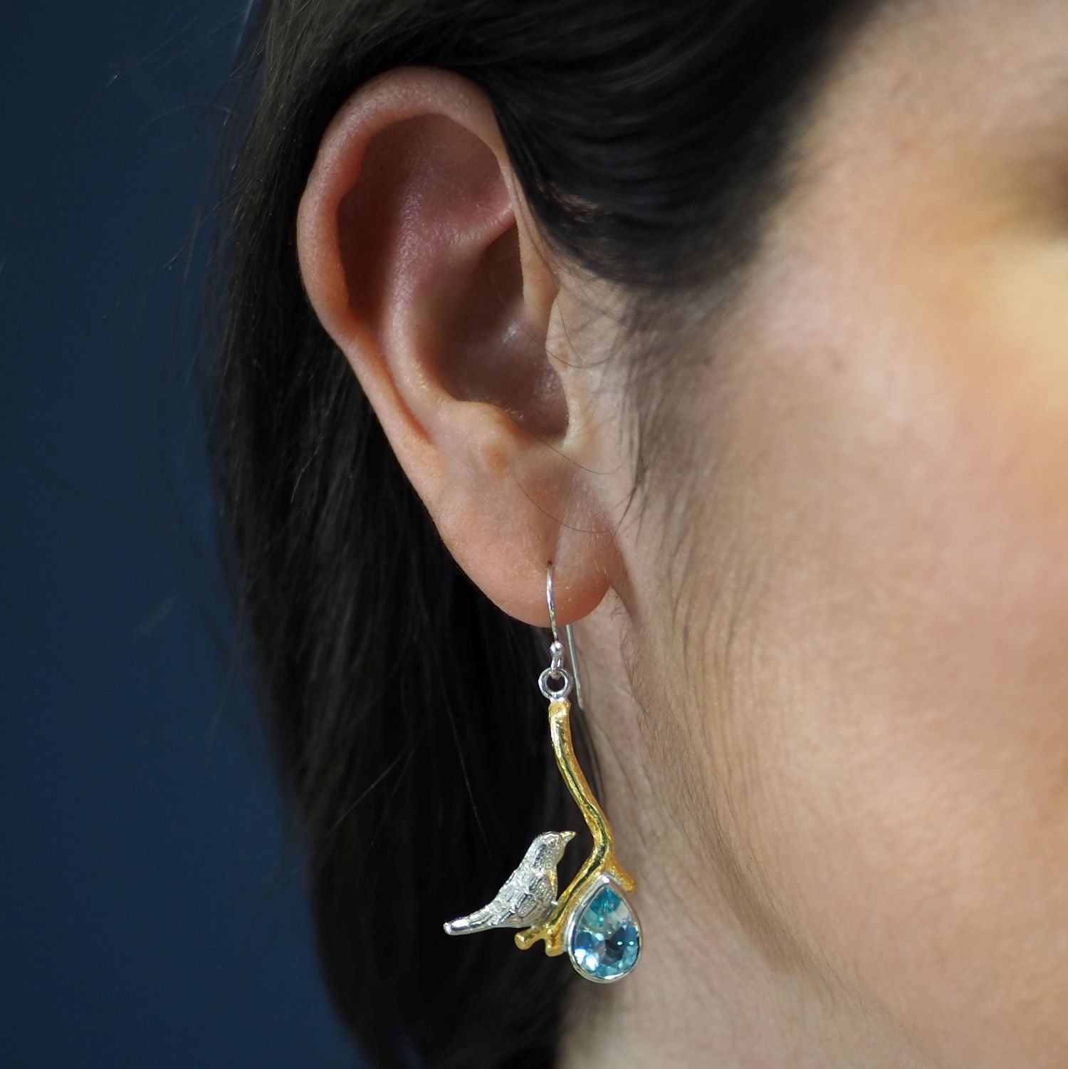 Close-up of a woman's ear wearing unique statement earrings. The earring features a small bird figure perched on a curved, gold-toned branch with a blue teardrop gemstone hanging below. The background is a simple, solid color. She is wearing the Lovebird 