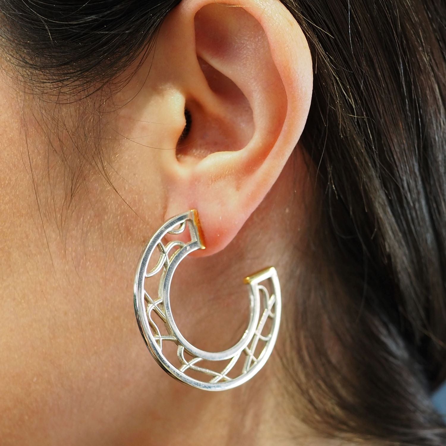 Close-up of a person's ear wearing a sterling silver hoop earring with an intricate, open lattice design. The Maya Earrings by Gallardo & Blaine Designs are shaped in a half-circle style and have a smooth, polished finish. The person's dark hair is partia