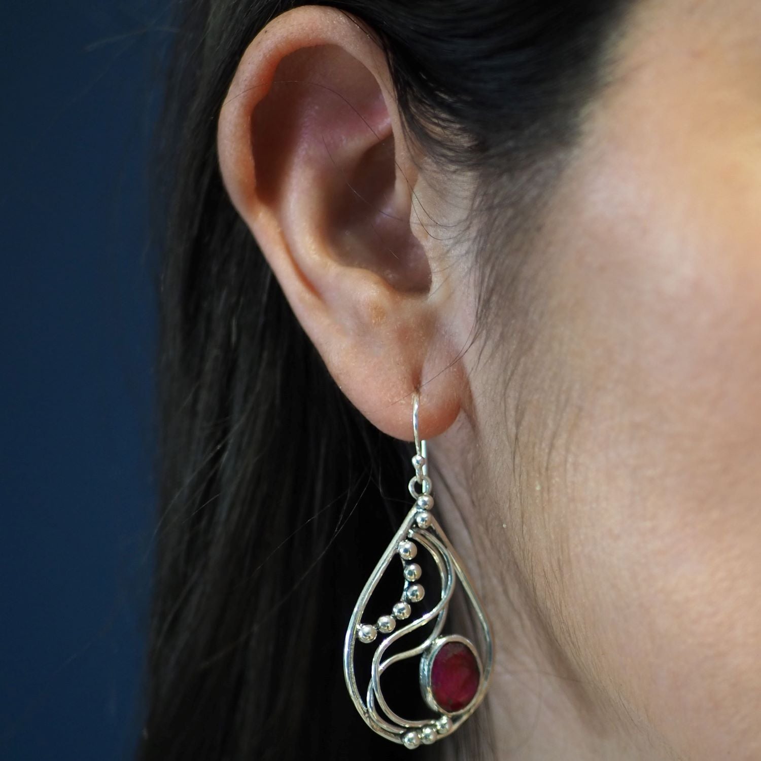Close-up of a woman's ear wearing a large, intricately designed Phoenix Earrings in various gemstones. The earring is silver with a teardrop shape, adorned with small spherical elements and a striking red oval gemstone near the bottom. The dark background