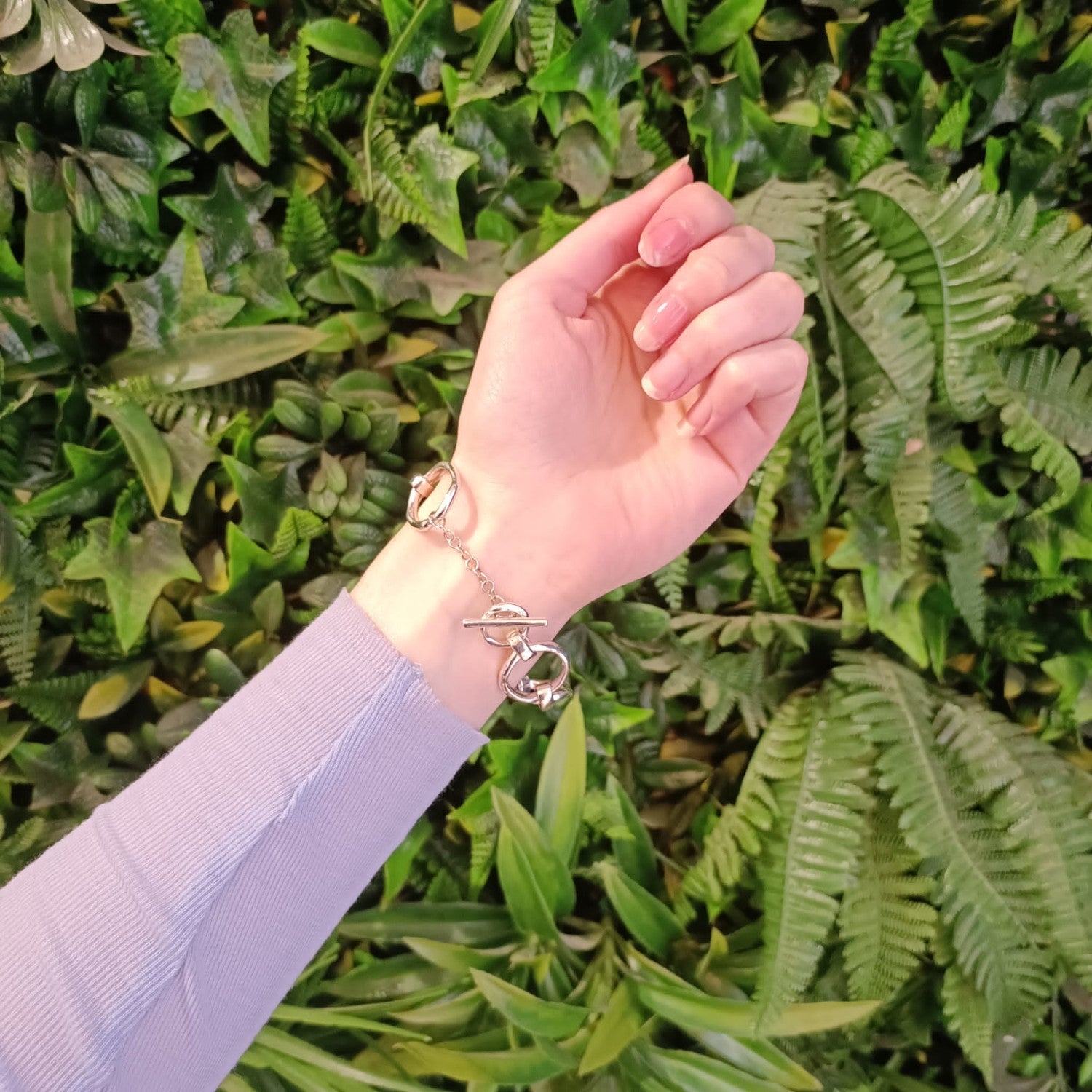 A hand wearing a Sea Honey Bracelet Silver by Gallardo & Blaine Designs featuring thick silver links and a clasp shaped like handcuffs is extended in front of a green leafy background. The arm is clothed in a long-sleeved, light purple top, showcasing the
