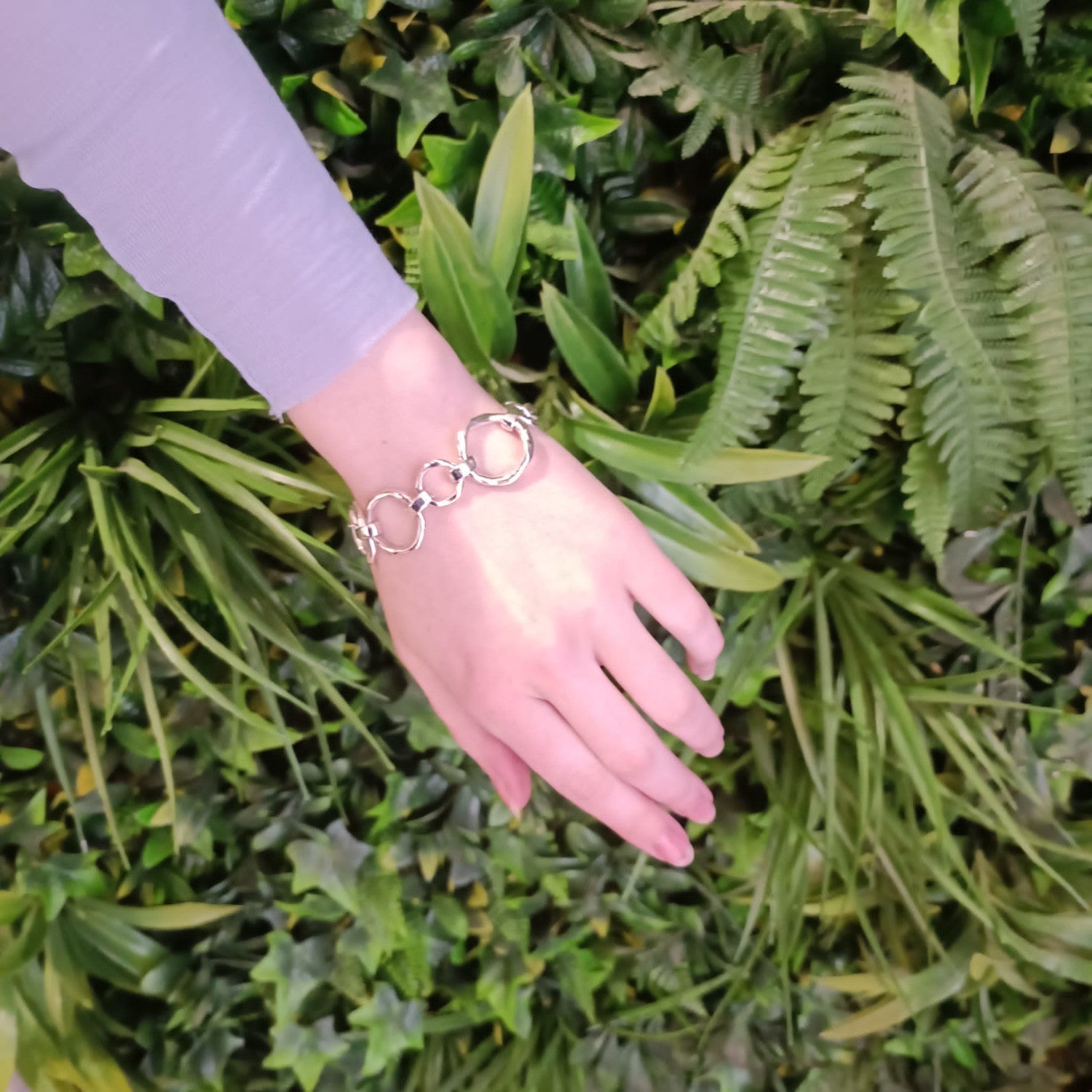 A hand with a Gallardo & Blaine Designs Sea Honey Bracelet Silver featuring thick silver links is extended against a background of lush green foliage. The person is wearing a long-sleeved, light purple top, showcasing one of the new designs.