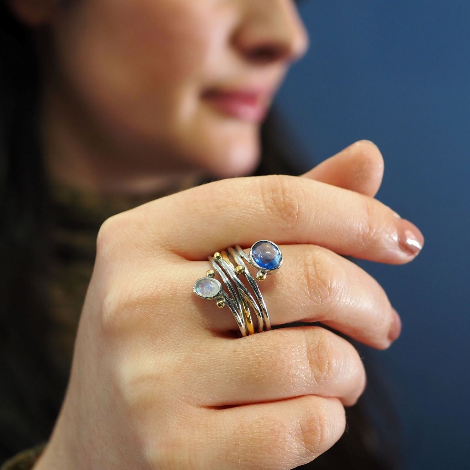 Close-up of a woman's hand wearing a stack of rings with blue gemstones, including a unique Gallardo & Blaine Designs Snake Ring in Silver Gold & various gemstones, held up to her face. The background is blurred, emphasizing the rings and the intricate de