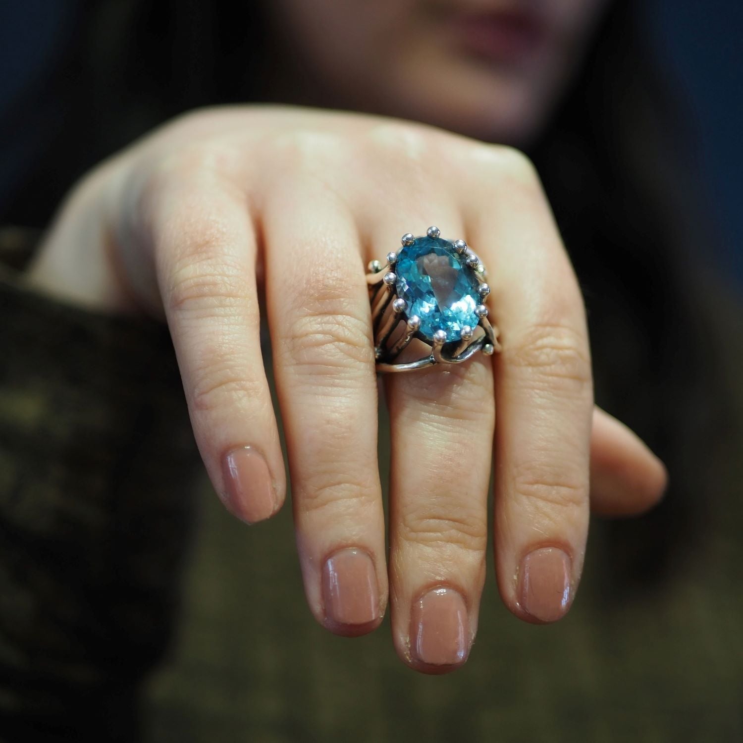 A close-up of a person's hand adorned with a chunky statement gemstone ring, the Willow Ring in various gemstones by Gallardo & Blaine Designs, featuring a prominent blue gemstone. The person's nails are neatly manicured with nude polish. The background i