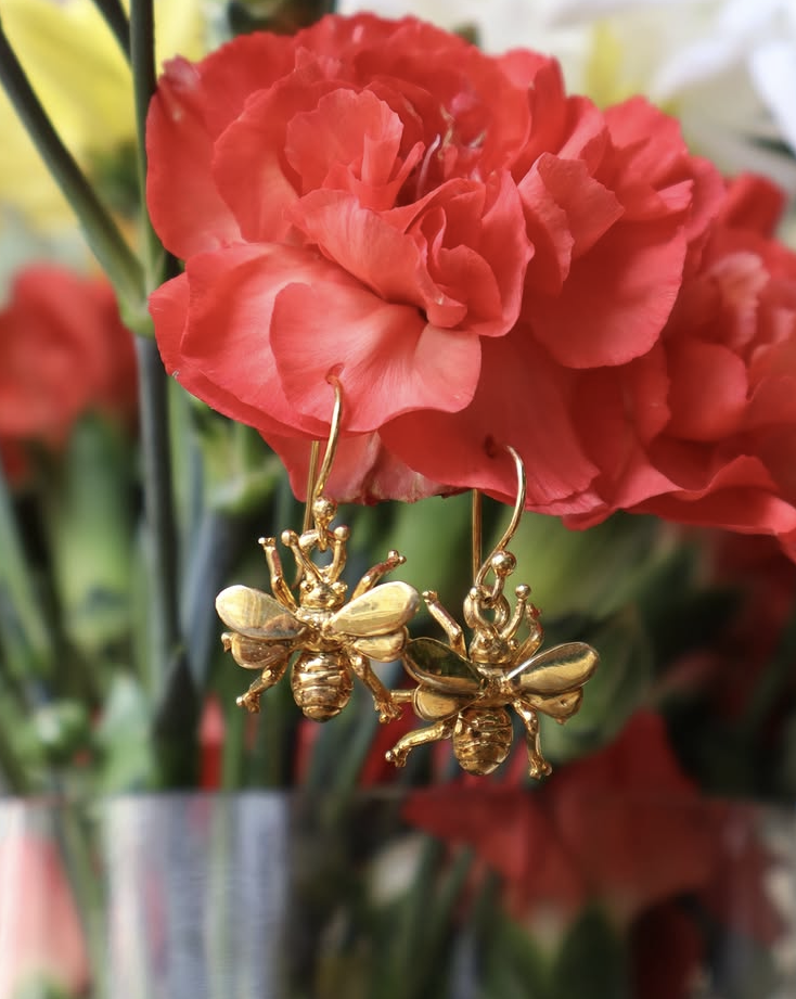 Watermelon Tropical’s Wildlife Bee Dangle Earrings (Silver or Gold) hang from the petals of a vivid red carnation, with blurred red and yellow blooms behind—an elegant nod to nature conservation–inspired wildlife jewelry.