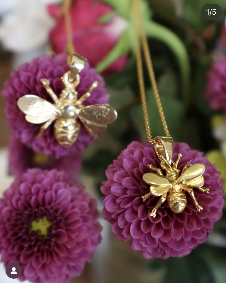 Two Wildlife Pendant - Large Bee With Chain necklaces by Watermelon Tropical are displayed on vibrant purple flowers with yellow-green centers, showcasing the charm of nature-inspired jewelry amidst vivid blooms.