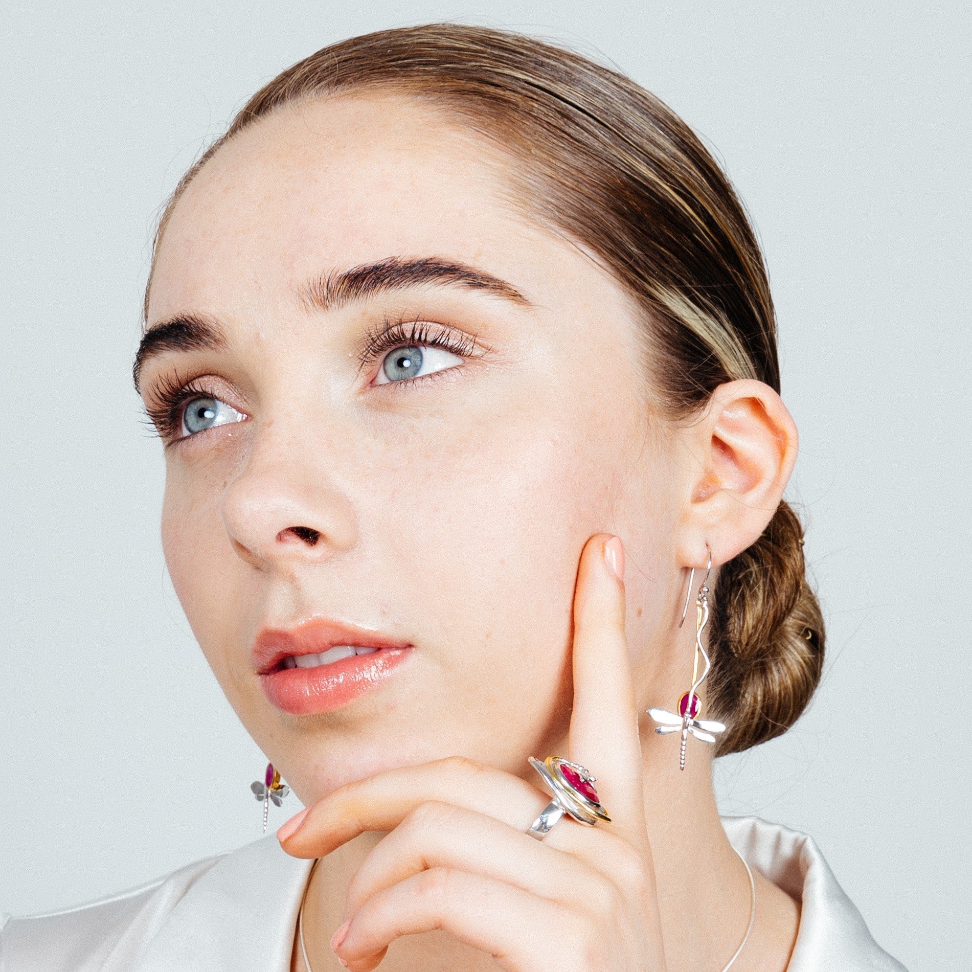 A contemplative person with slicked-back hair wearing a white shirt, Willow Earrings in Various Gemstones by Gallardo & Blaine Designs, and a matching ring with a gemstone. They are resting their chin on their fingers, gazing upward with a serene expressi