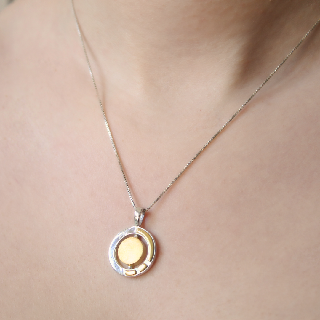 A close-up of a person’s neck wearing the Urú Small Pendant & Chain by Gallardo & Blaine Designs, featuring a modern, minimalist round pendant that elegantly blends sterling silver and gold tones.